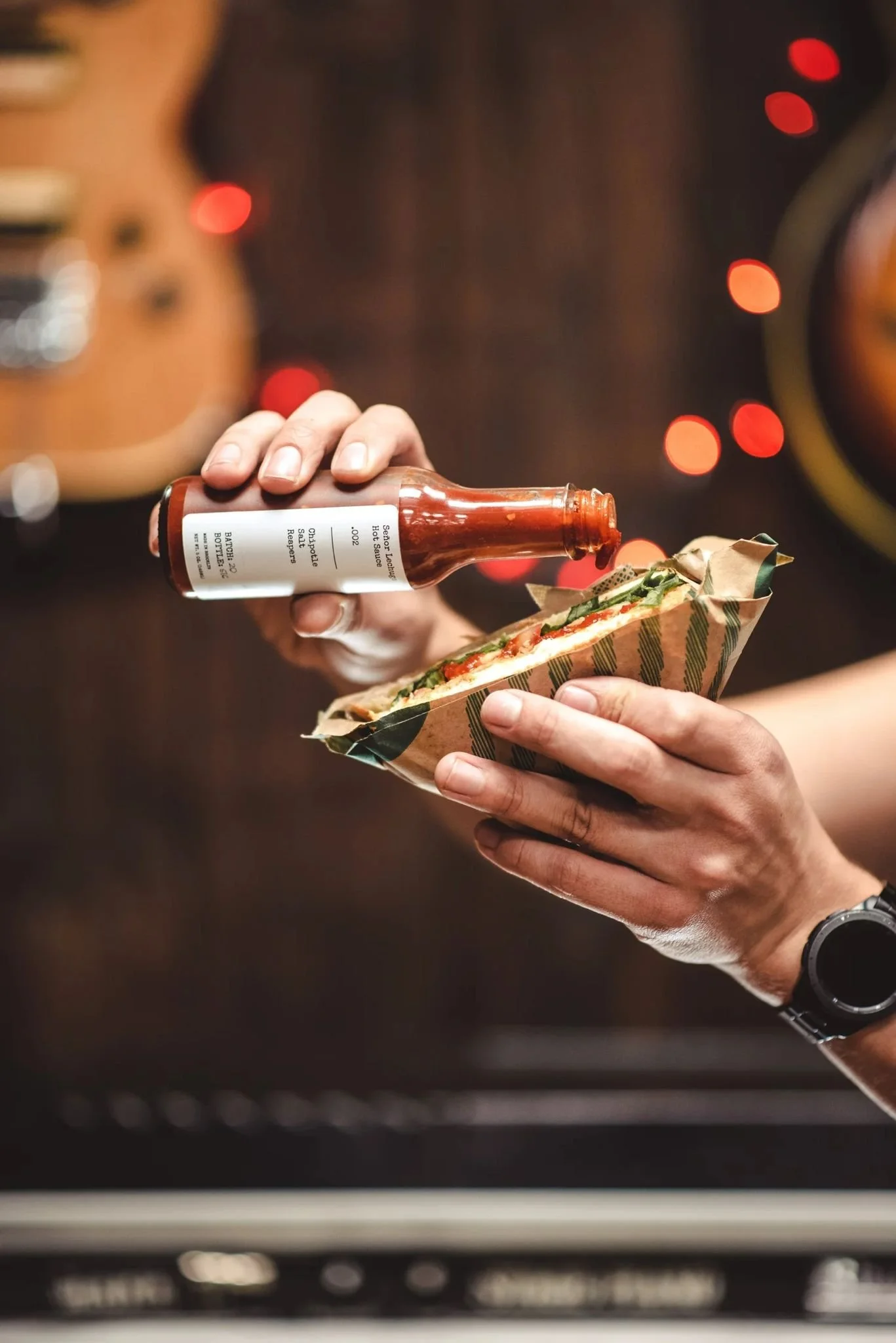 Person holding a sandwich with a bottle of hot sauce above it, in a setting with red bokeh lights in the background.