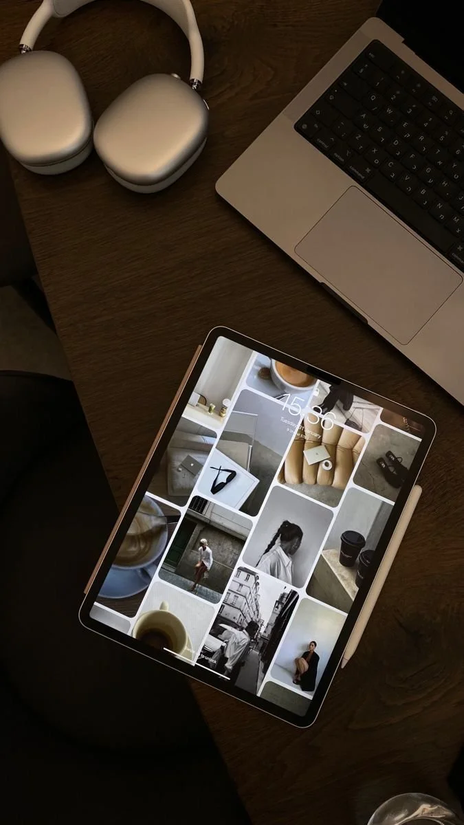 A wooden table with a pair of white over-ear headphones, a silver laptop, and a tablet displaying a collage of black-and-white and color photos of people and objects.