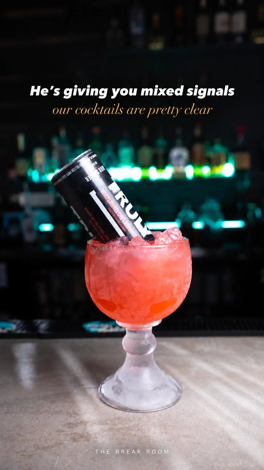 A pink cocktail with ice in a glass goblet, with a black energy drink can inserted into it, against a bar background with illuminated bottles.