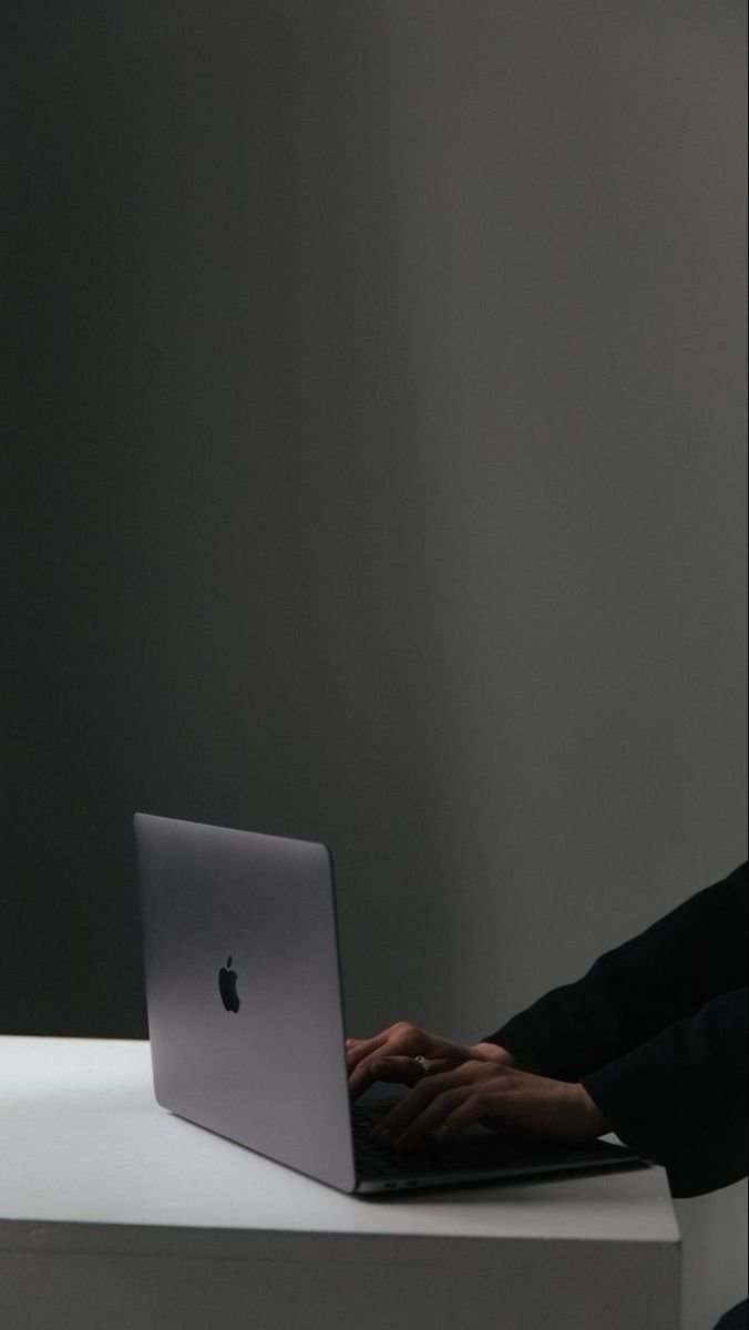 Person in black clothing typing on a silver MacBook laptop placed on a white table in a dimly lit room.