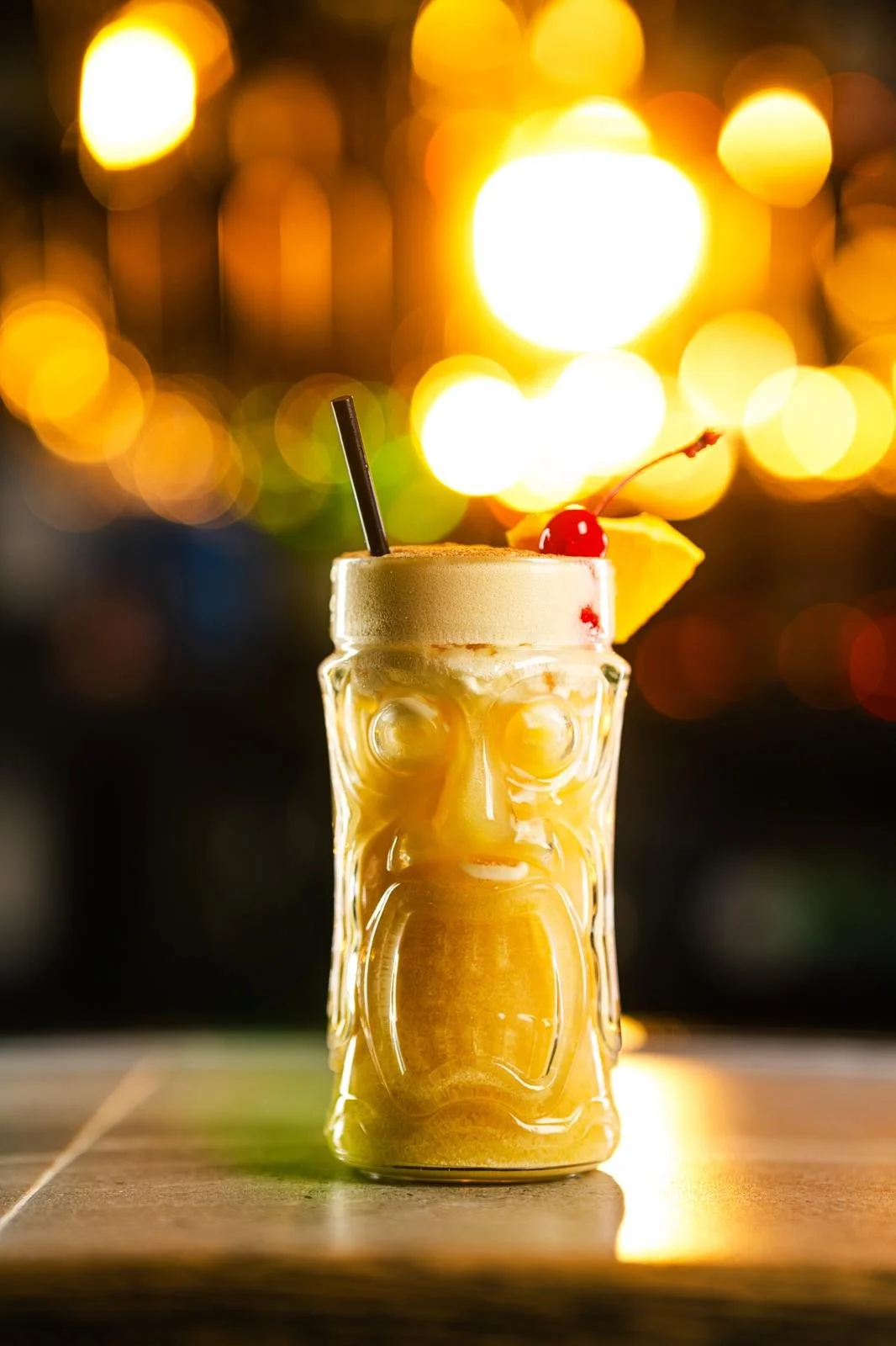 A tiki-style glass filled with a yellow tropical cocktail, garnished with a cherry and pineapple, with a straw, on a wooden surface at sunset or evening with blurred city lights in the background.