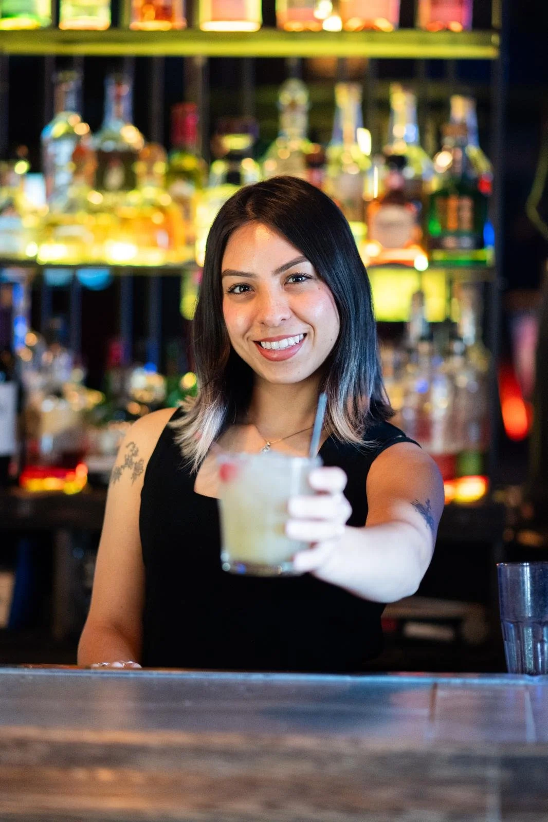 A woman with black hair and tattoos holding a cocktail in a bar with colorful bottles in the background.