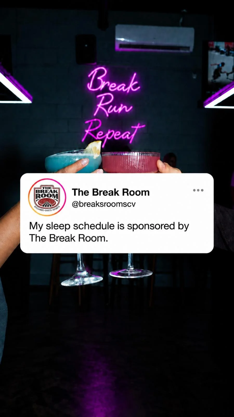 Two hands holding cocktails with a neon sign reading 'Break Run Repeat' in the background at The Break Room bar.