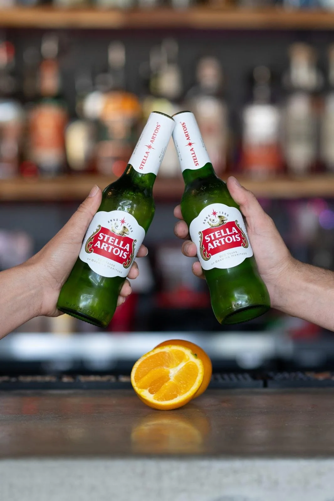 Two people holding Stella Artois beer bottles clinking together with orange half on bar counter below.