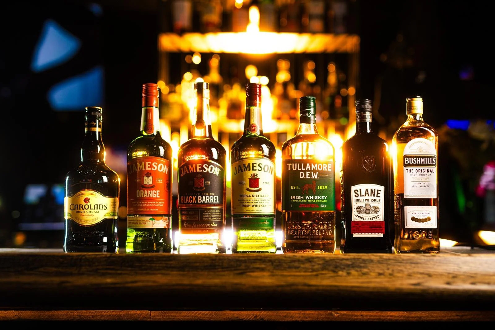Six bottles of Irish whiskey and liqueur on a bar counter with blurred bottles and warm lighting in the background.