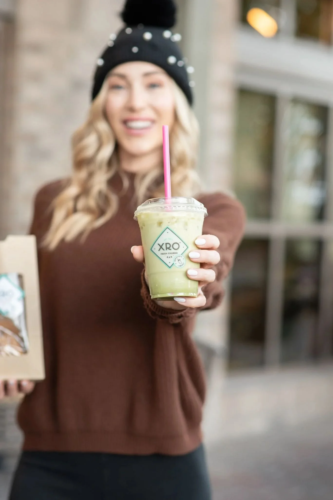 A woman holding a green drink in a clear plastic cup with a pink straw, smiling and wearing a black beanie with pearl embellishments.