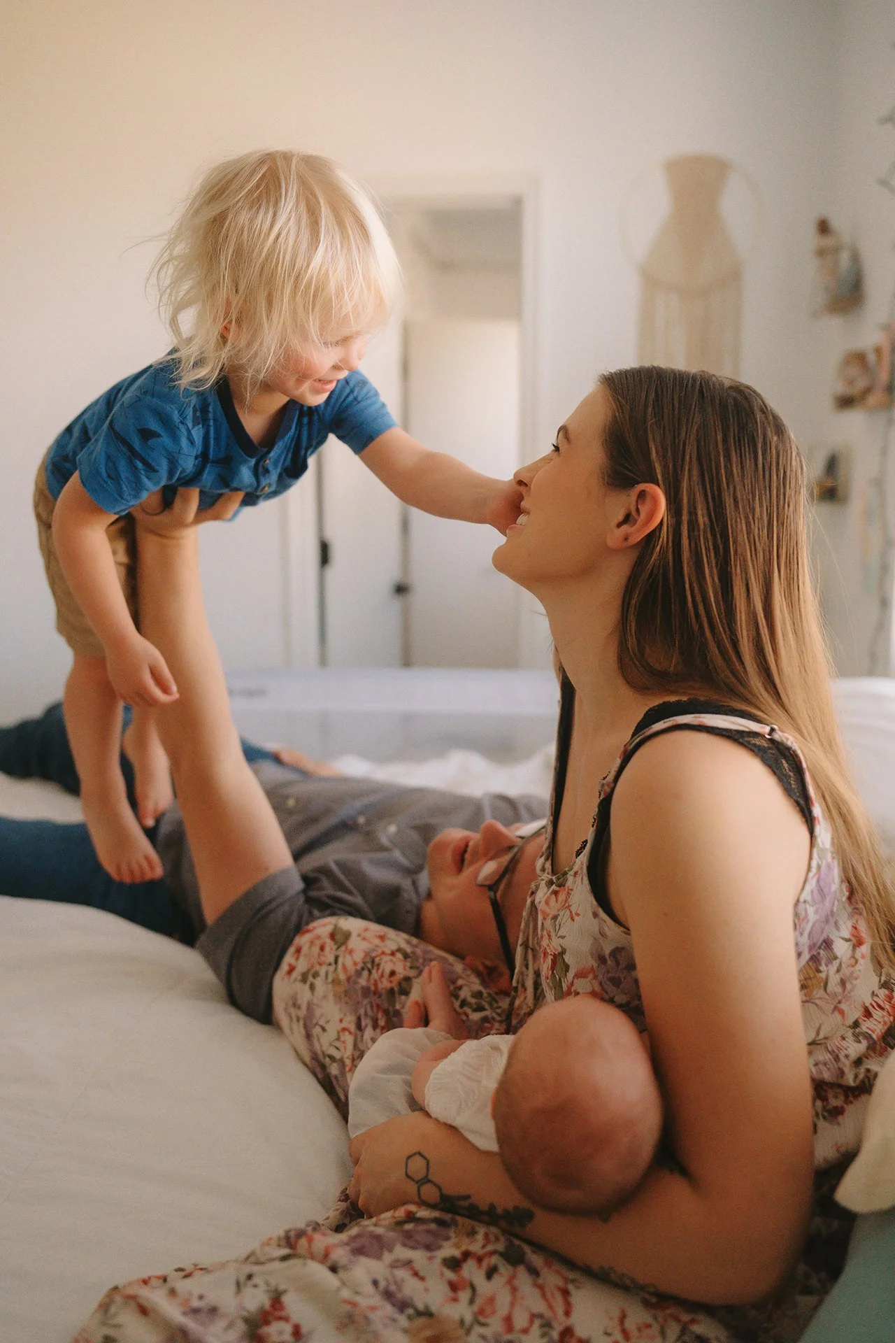 A playful portrait of a father laying down on his bed, holding his blonde-haired toddler at eye level with his beautiful brunette wife, who sits beside them, cradling their newborn daughter.