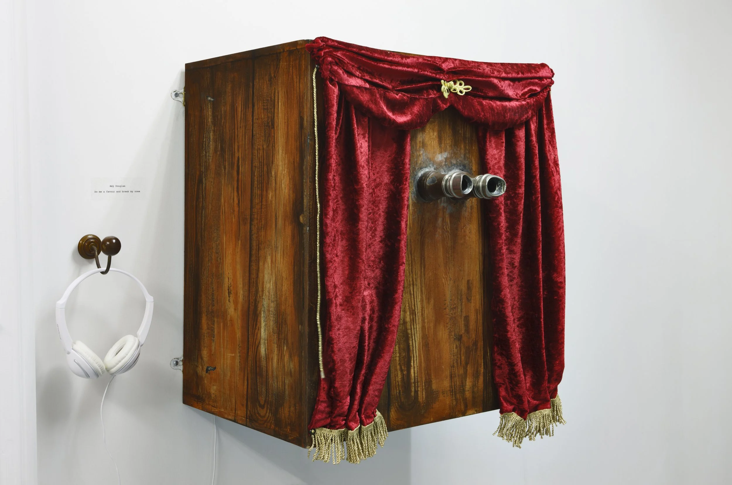 Antique wooden viewing box with red velvet curtains and binocular-like lenses, mounted on a wall beside white headphones on a hook.