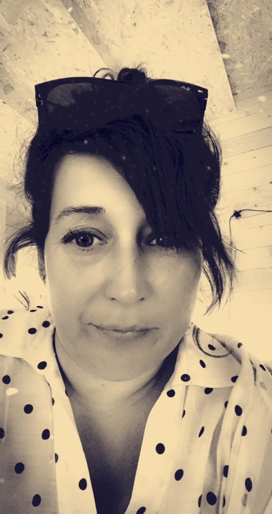 Black and white selfie of a woman with short dark hair, sunglasses on her head, and a polka dot blouse, smiling slightly, indoors with a wooden and textured wall background.