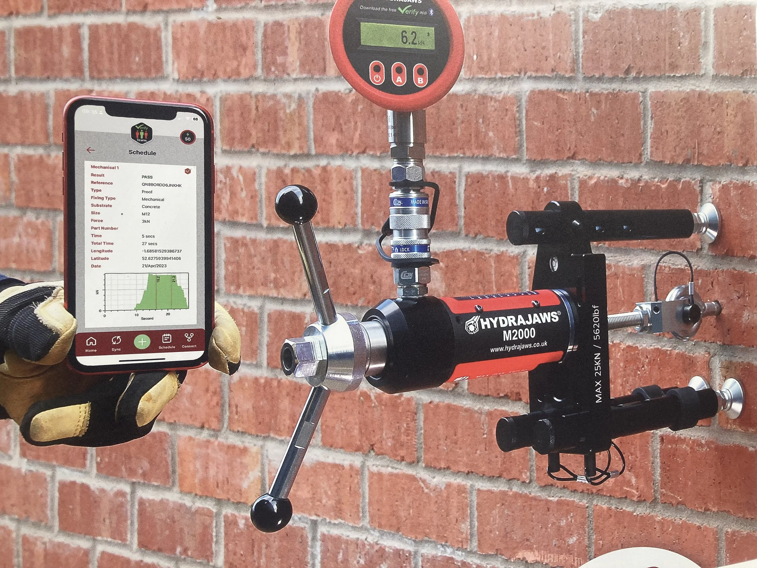 A hand holding a smartphone with a testing app on the screen, next to a piece of laboratory equipment mounted on a brick wall, connected to a digital force gauge showing 6.2 kN.