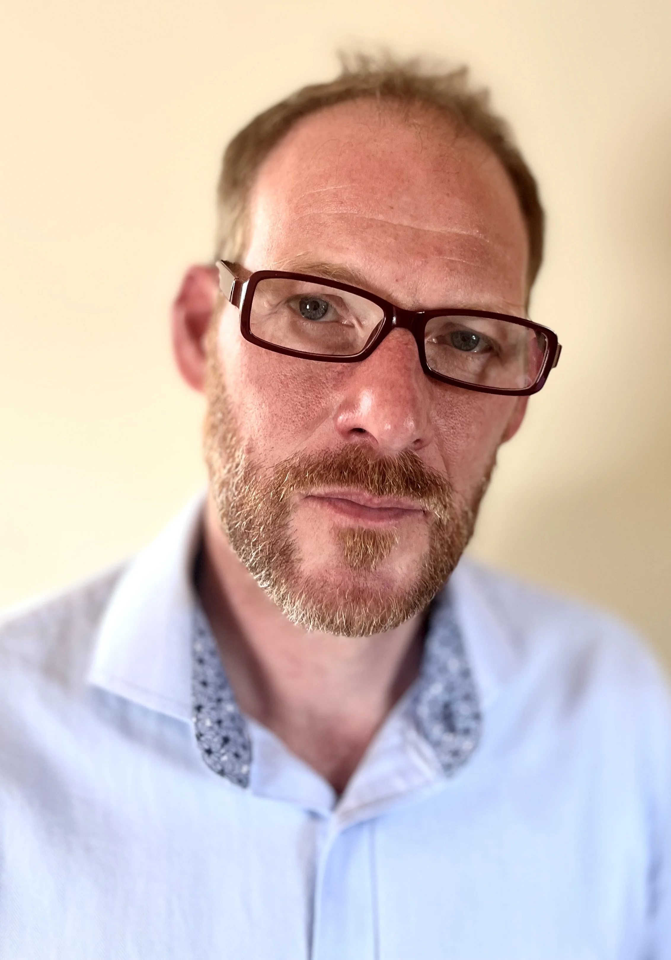 Close-up of a man with glasses, reddish beard, and short hair, wearing a light blue shirt with a patterned collar, standing against a plain light-colored background.