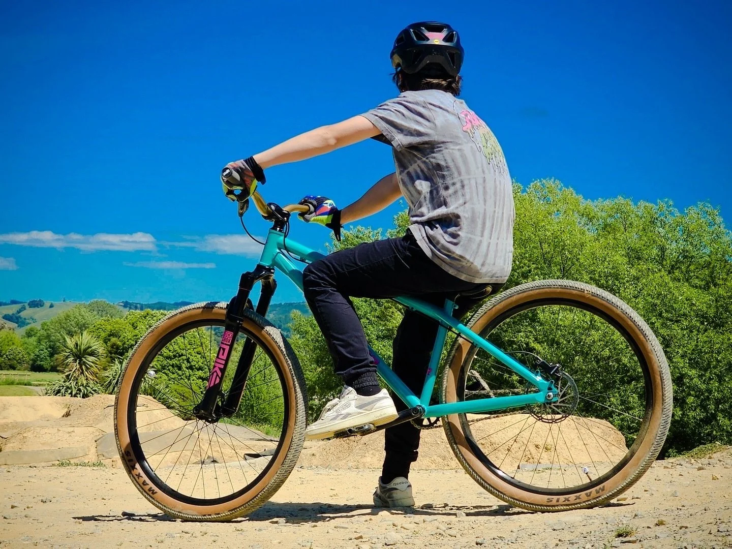 A child riding a teal mountain bike on a dirt path in a scenic outdoor area with green trees and hills under a bright blue sky.
