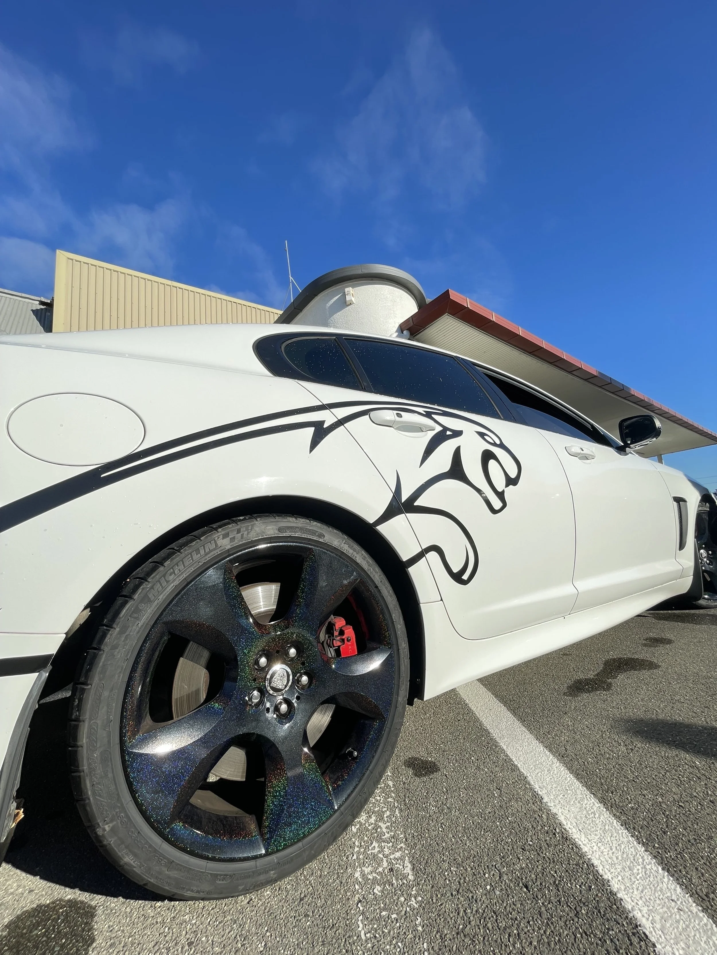 Close-up of a white sports car with a black infographic of a panther on the side, parked in a lot under a clear blue sky.