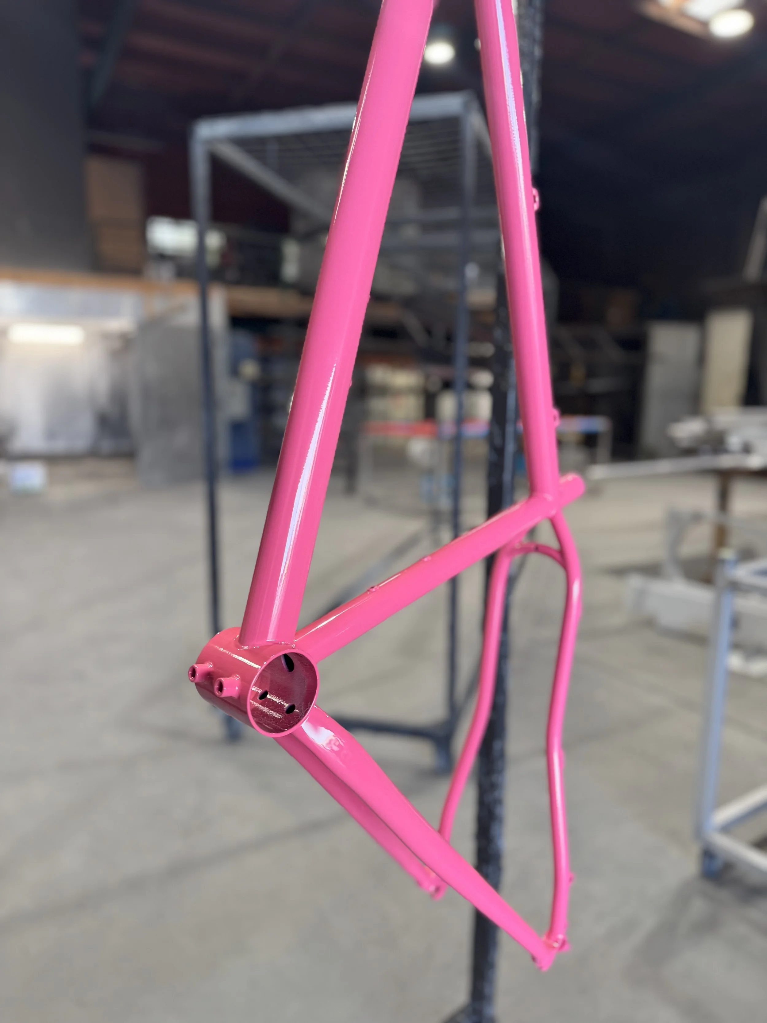 Close-up view of a pink bicycle frame hanging on a metal rack in a warehouse or workshop setting, with shelving and other items in the background.