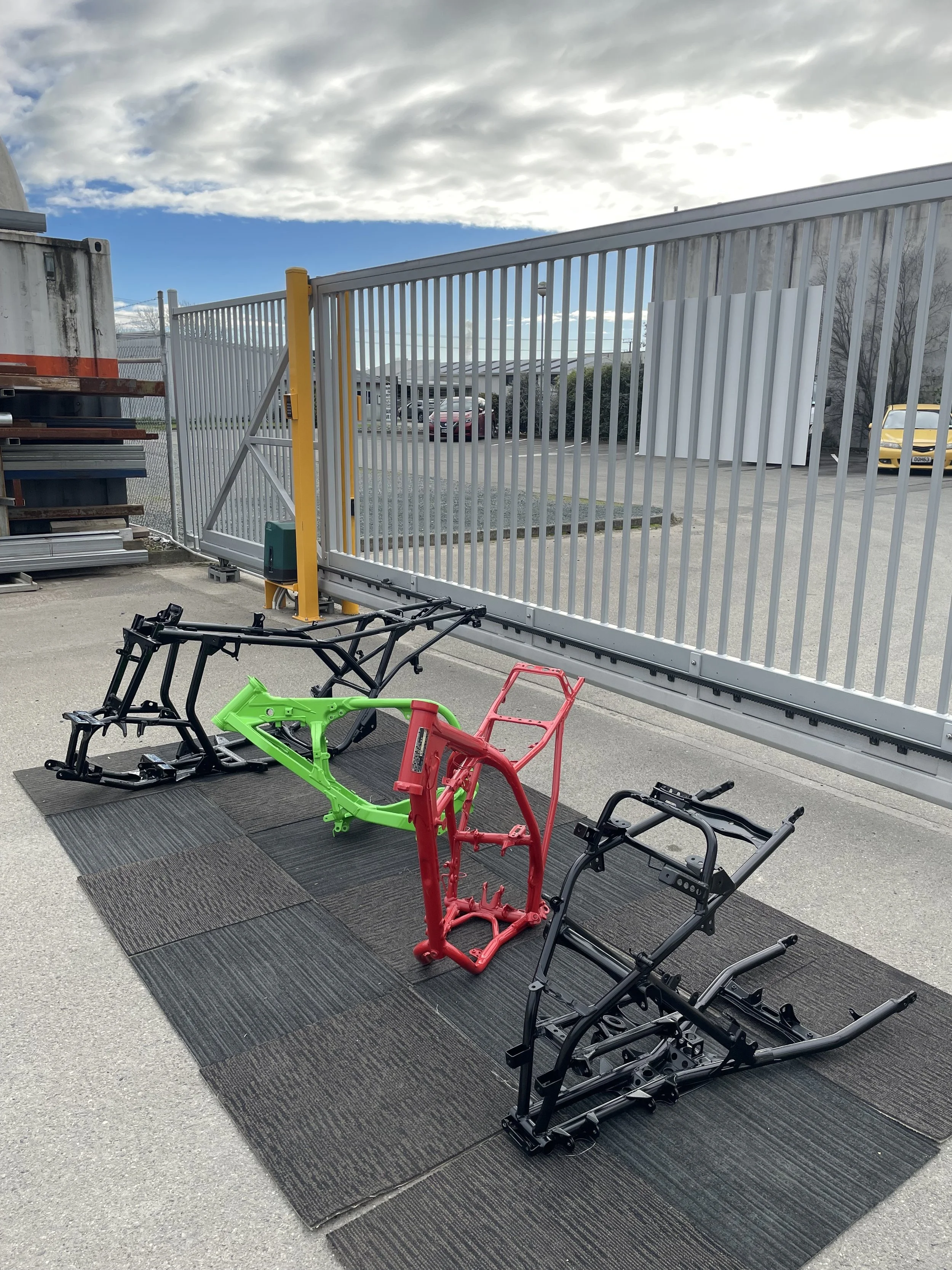 Colorful bicycle frames displayed on a mat outdoors with a metal fence, parking lot, cars, and a partly cloudy sky in the background.