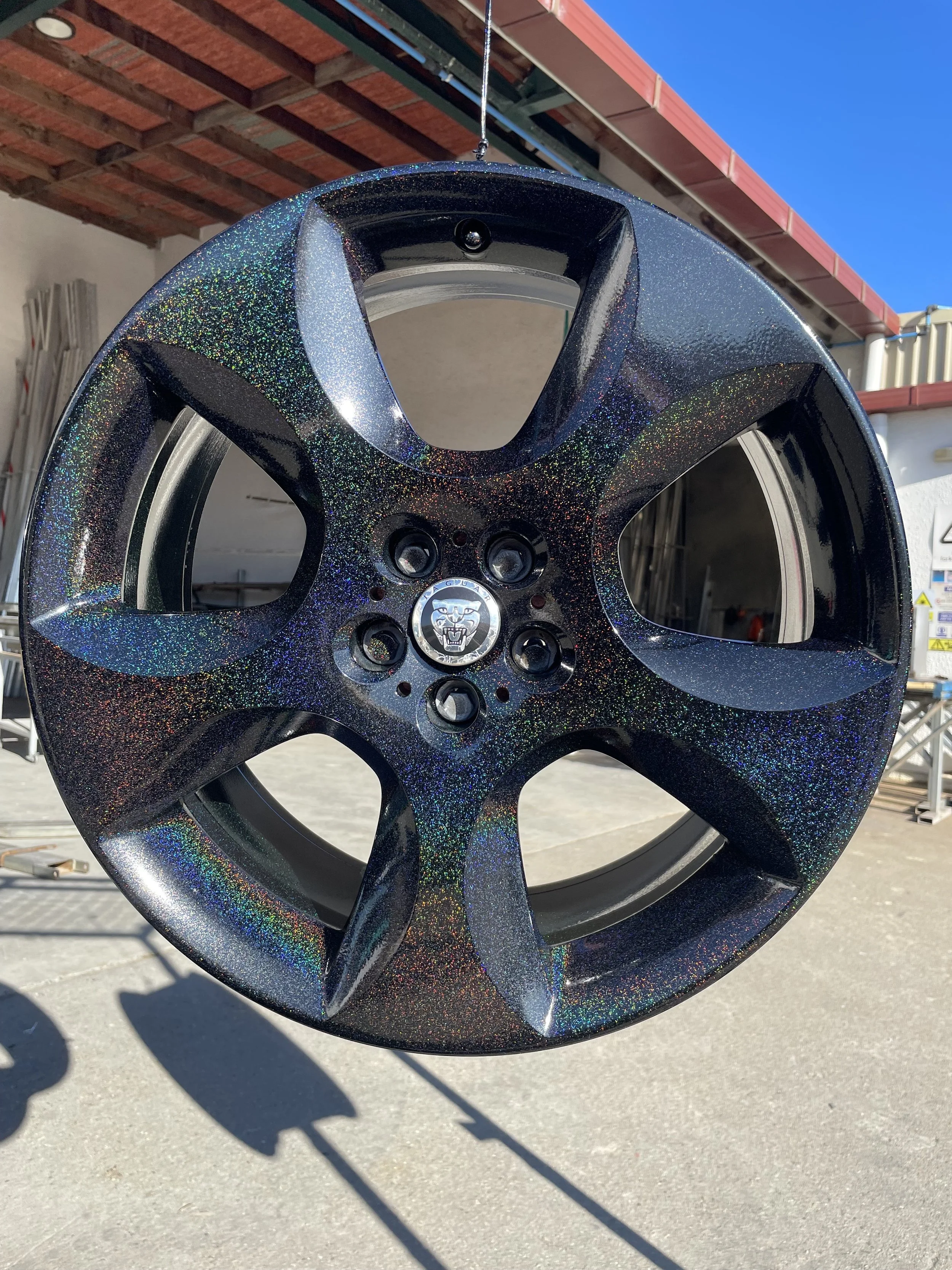 Car alloy wheel with a black and holographic glitter finish, hanging from a thin wire outdoors with a sunny sky and building in the background.