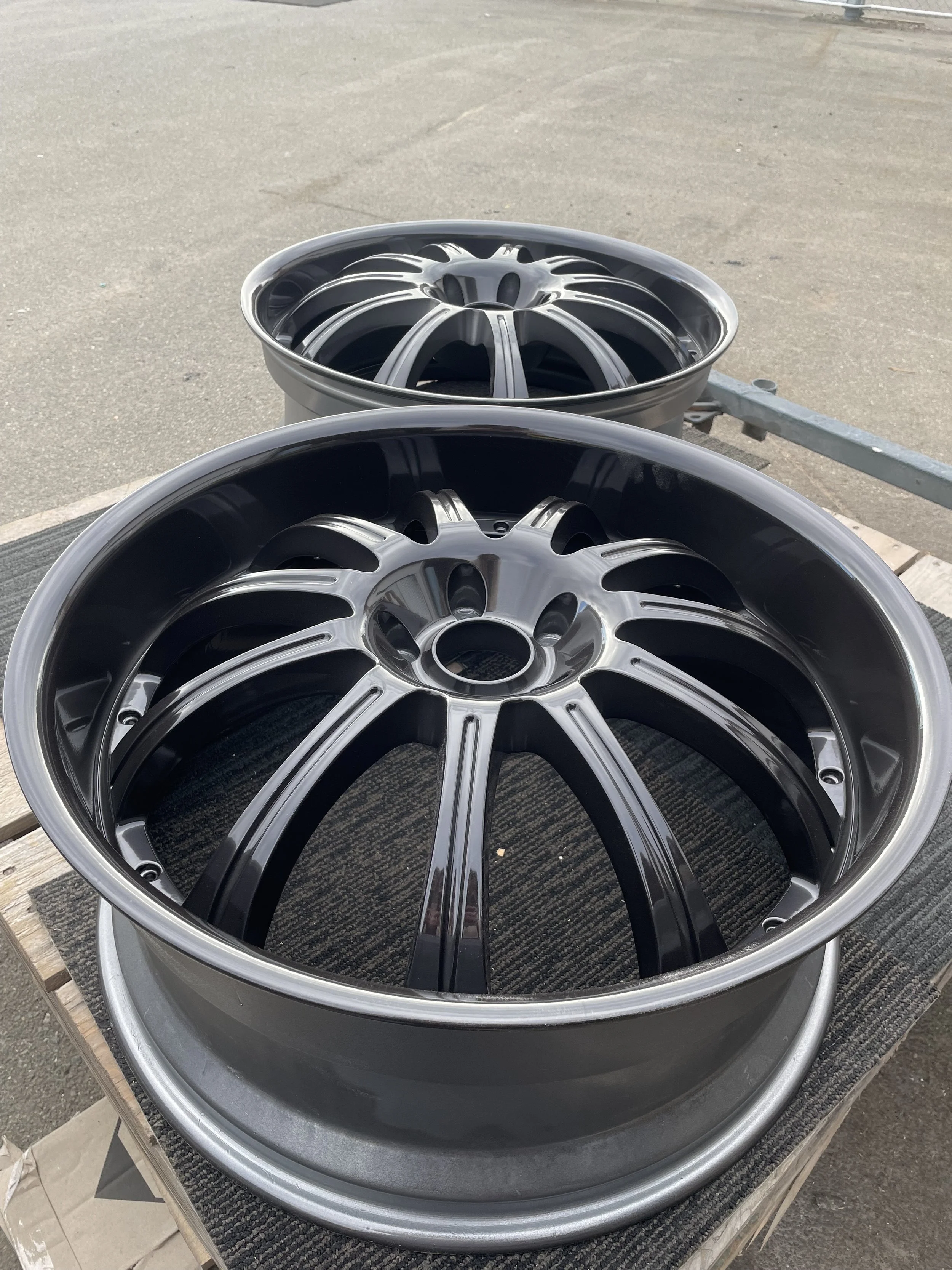 Two black and silver car wheels with intricate spoke designs placed outdoors on a wooden surface.
