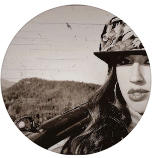 Close-up black-and-white photo of a woman wearing a hat with palm leaf pattern, outdoors with a background of hills and sky.
