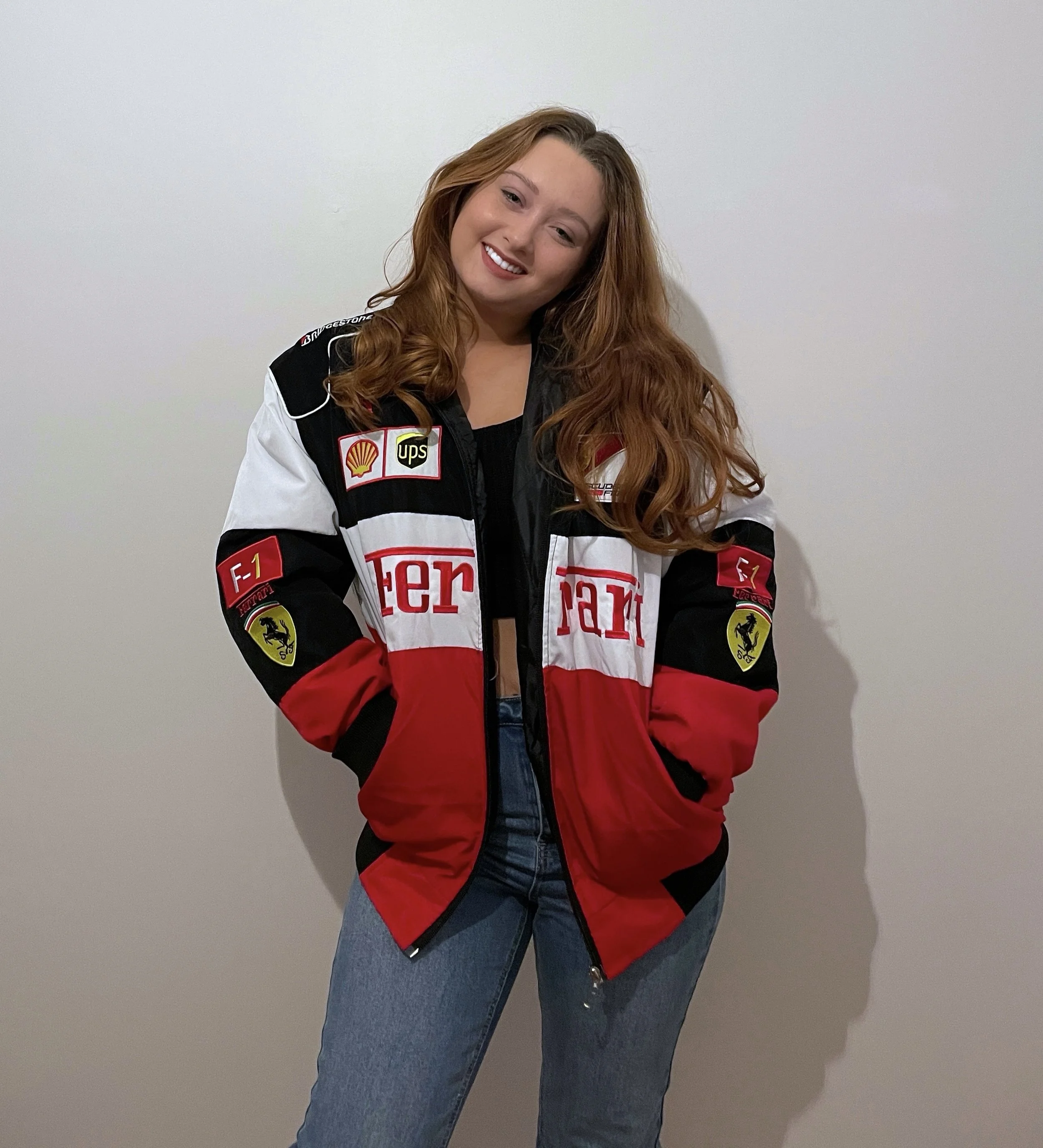 A young woman with long, wavy red hair, smiling and standing against a plain white wall, wearing a racing jacket with Ferrari logos and patches, and blue jeans.
