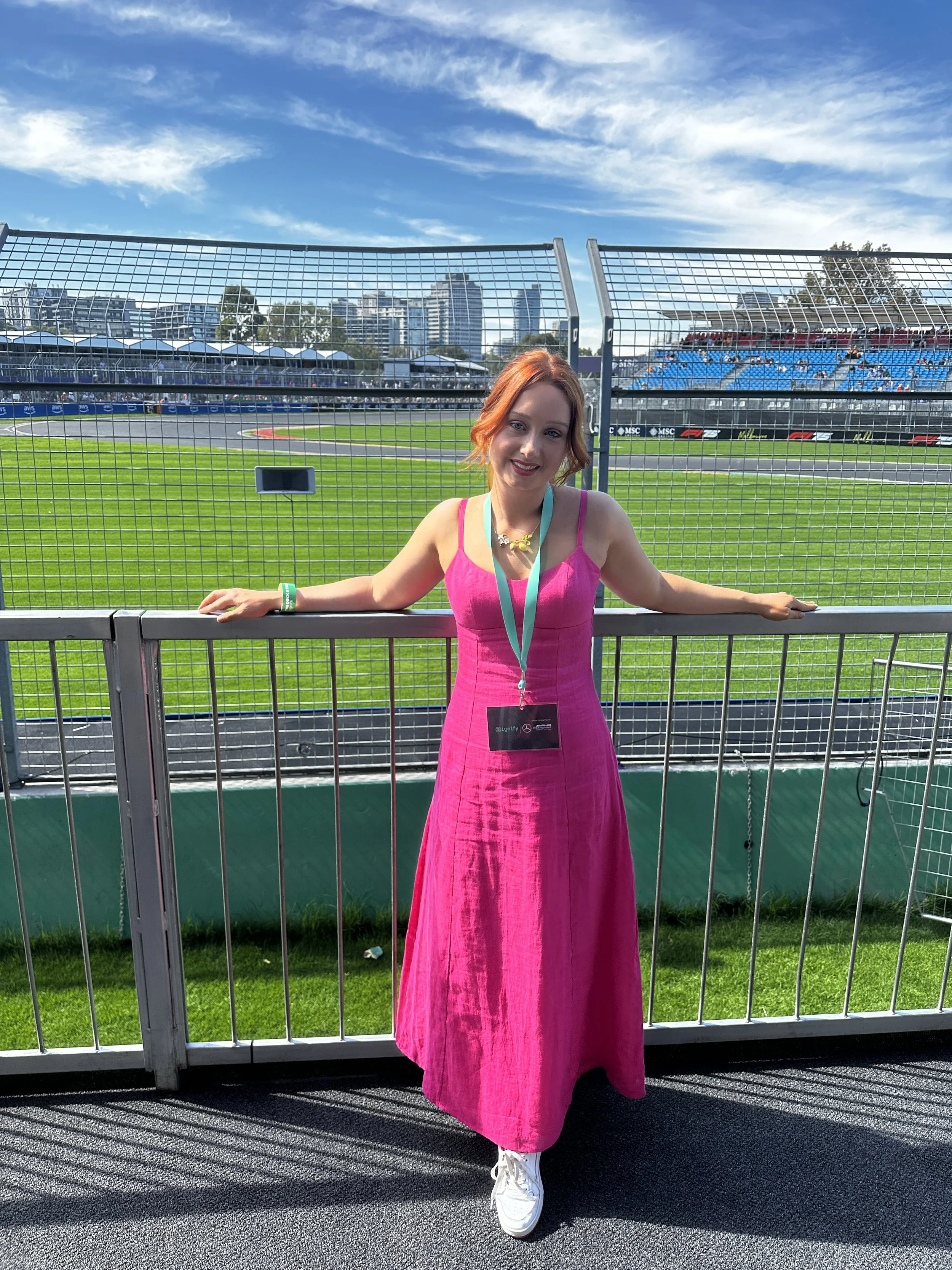 A woman with red hair in a pink dress standing by a metal fence at a race track with a city skyline and blue sky in the background. Australian Grand Prix Hospitality 2025.