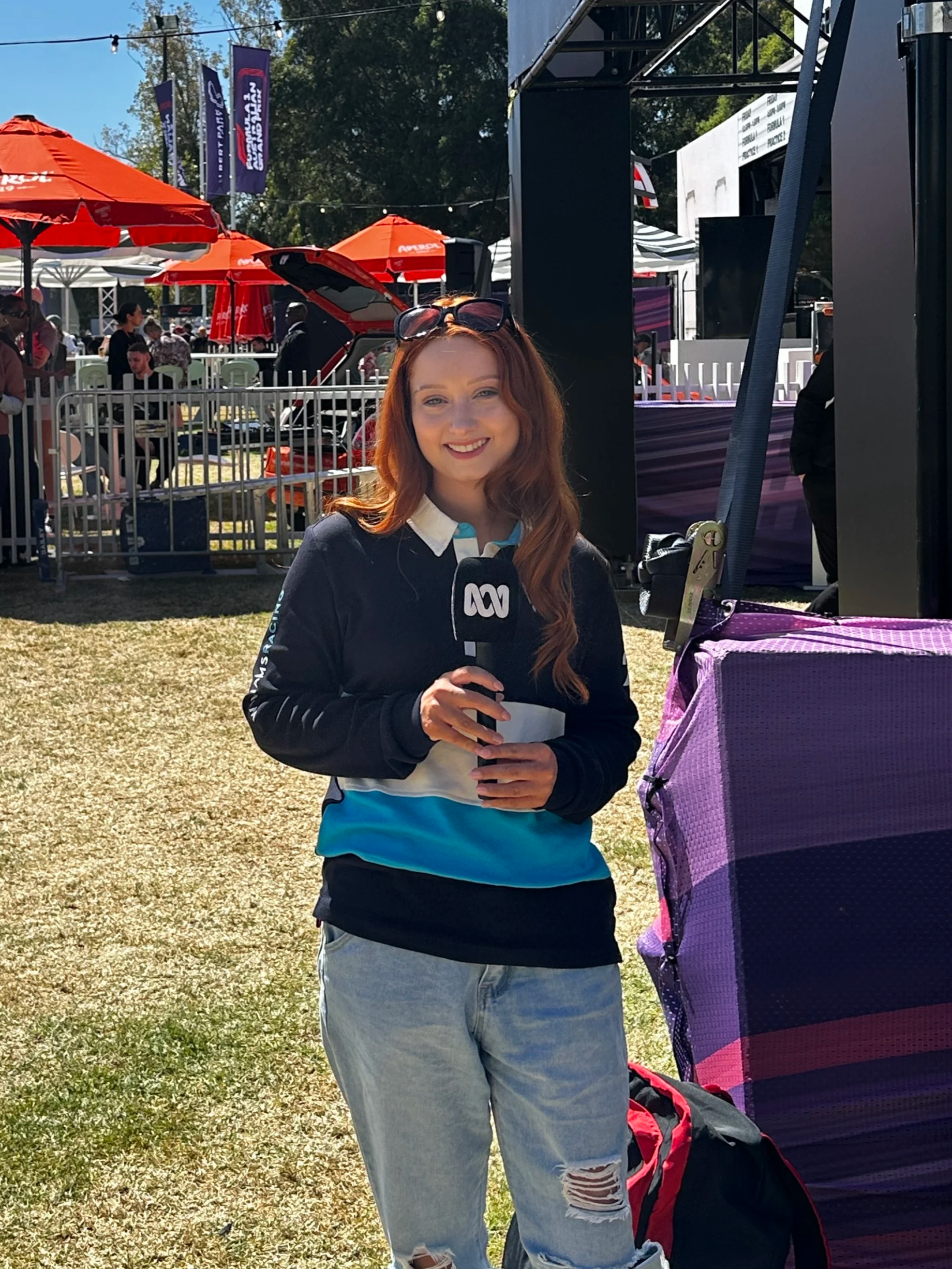 A female reporter with long red hair holds an ABC microphone at an outdoor event on a sunny day, with people, umbrellas, and tents in the background.