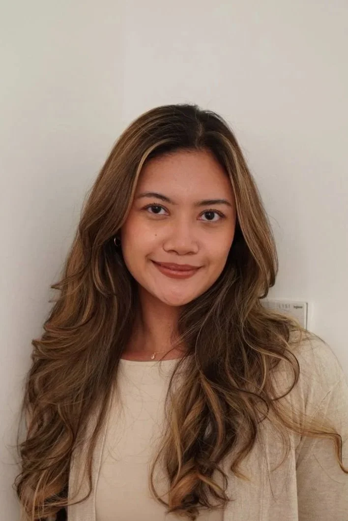 A young woman with long, wavy light brown hair, wearing a beige top and small hoop earrings, smiling softly in front of a plain off-white wall.