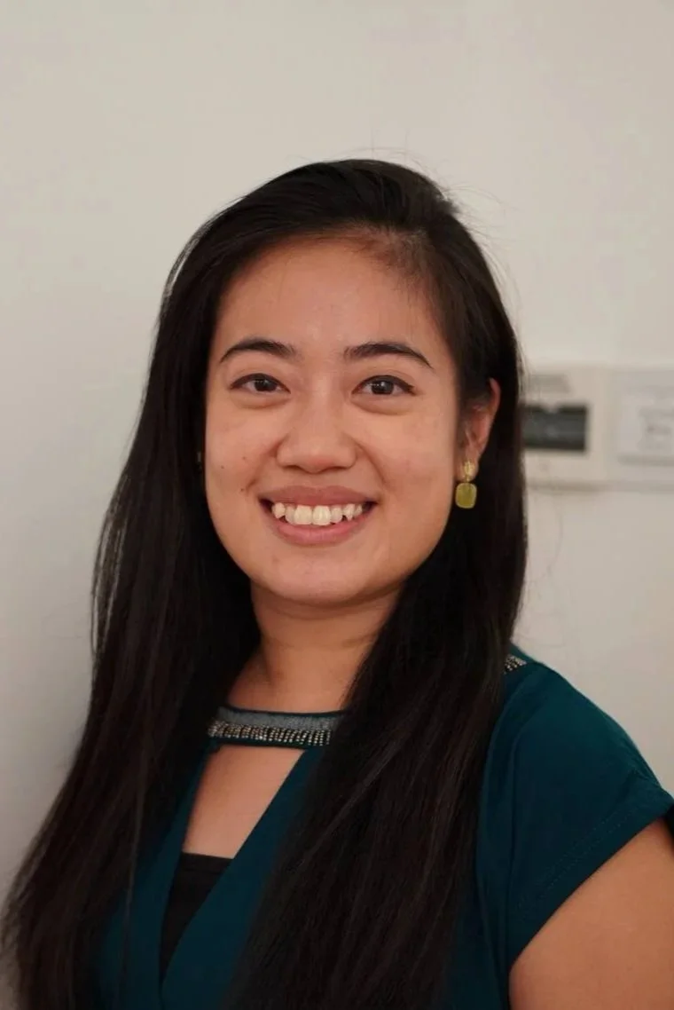 A woman with long dark hair smiling, wearing a teal top with embellishments near the neckline and gold earrings, standing in front of a white wall with electrical panels in the background.