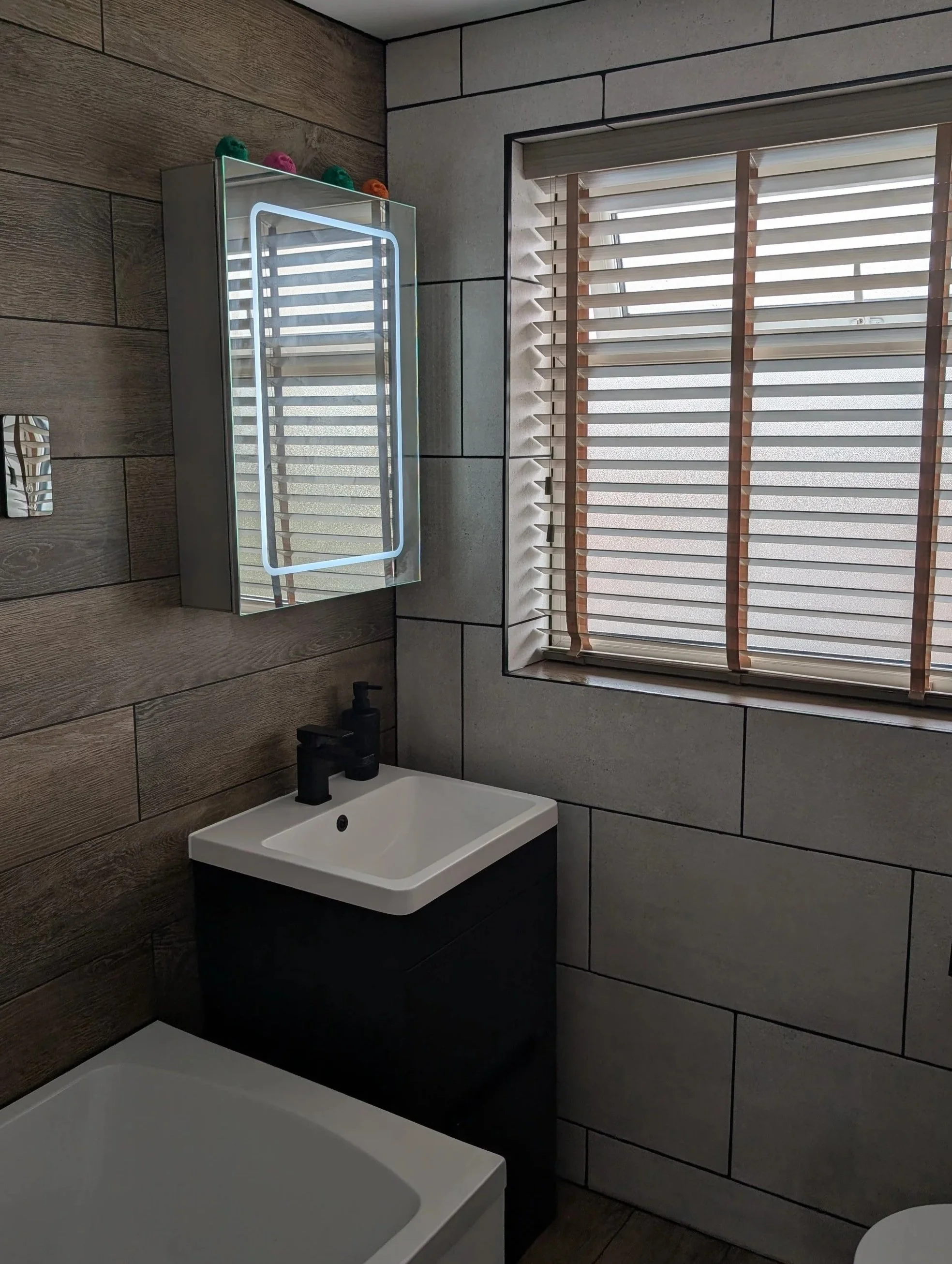 Small modern bathroom with a rectangular mirror with LED lighting, a white sink with black fixtures, a window with brown blinds, and wooden and gray tiled walls.