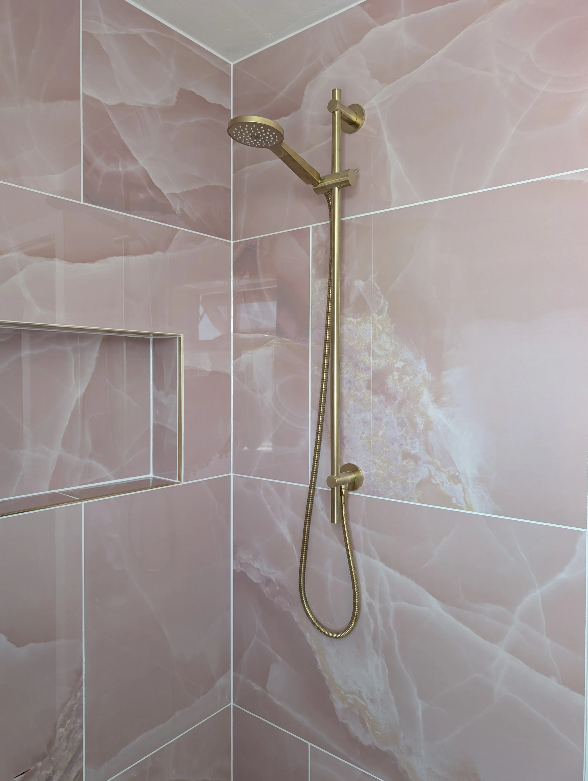 A shower wall with pink marble tiles, a built-in square shelf, and a gold-colored showerhead with a flexible hose.