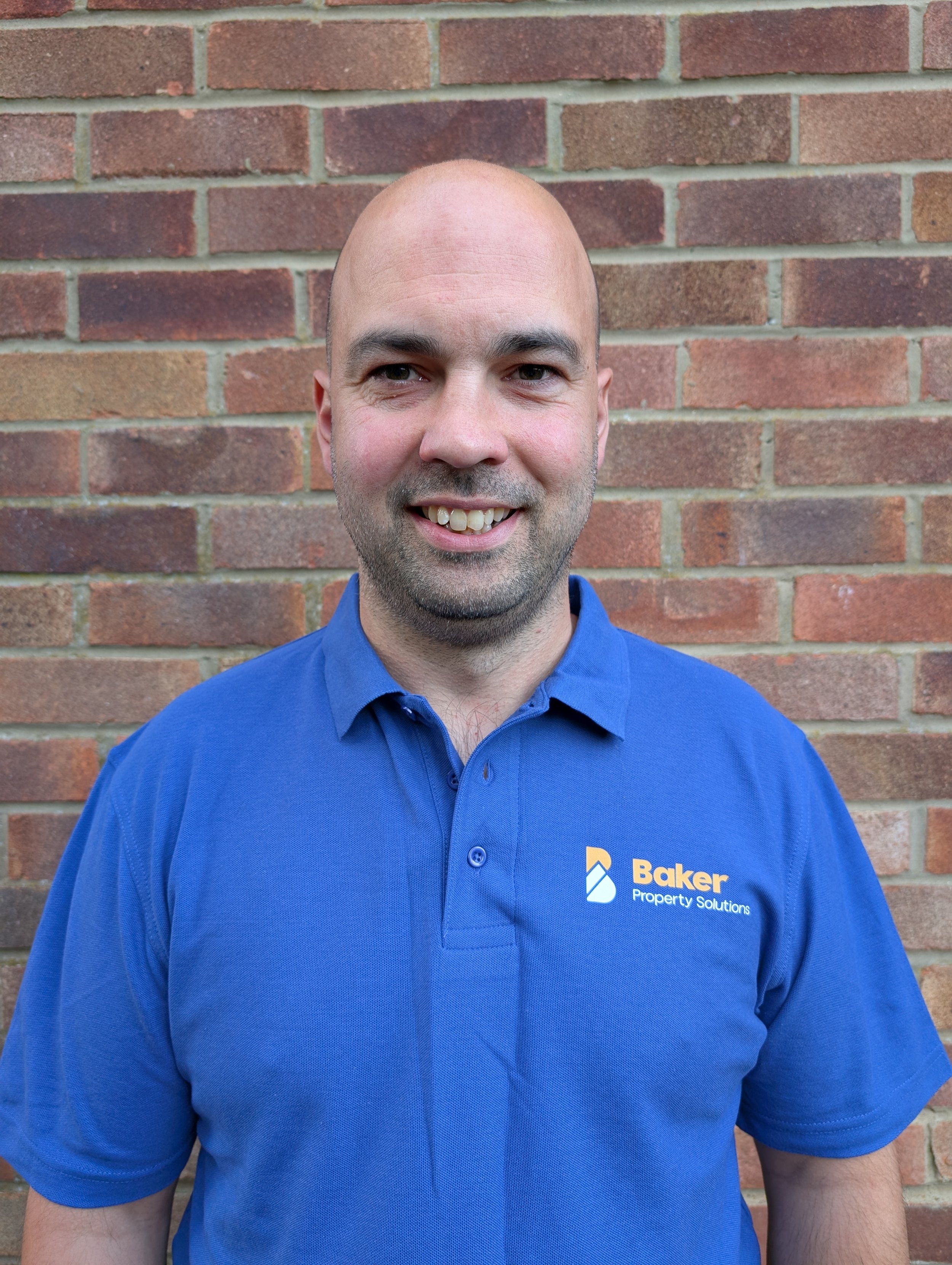 A smiling man wearing a blue polo shirt with Baker Property Solutions logo, standing in front of a brick wall.