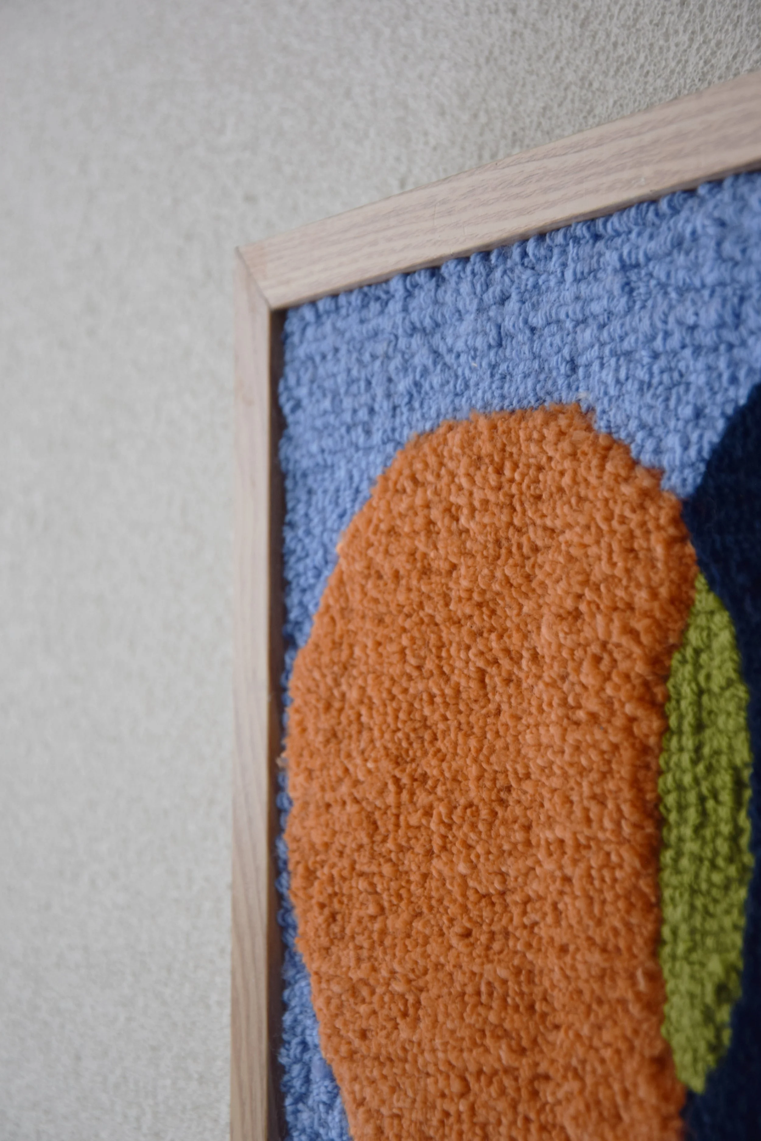 Close-up of textile wall art with colorful, textured patterns, framed with light wood, mounted on a beige wall.