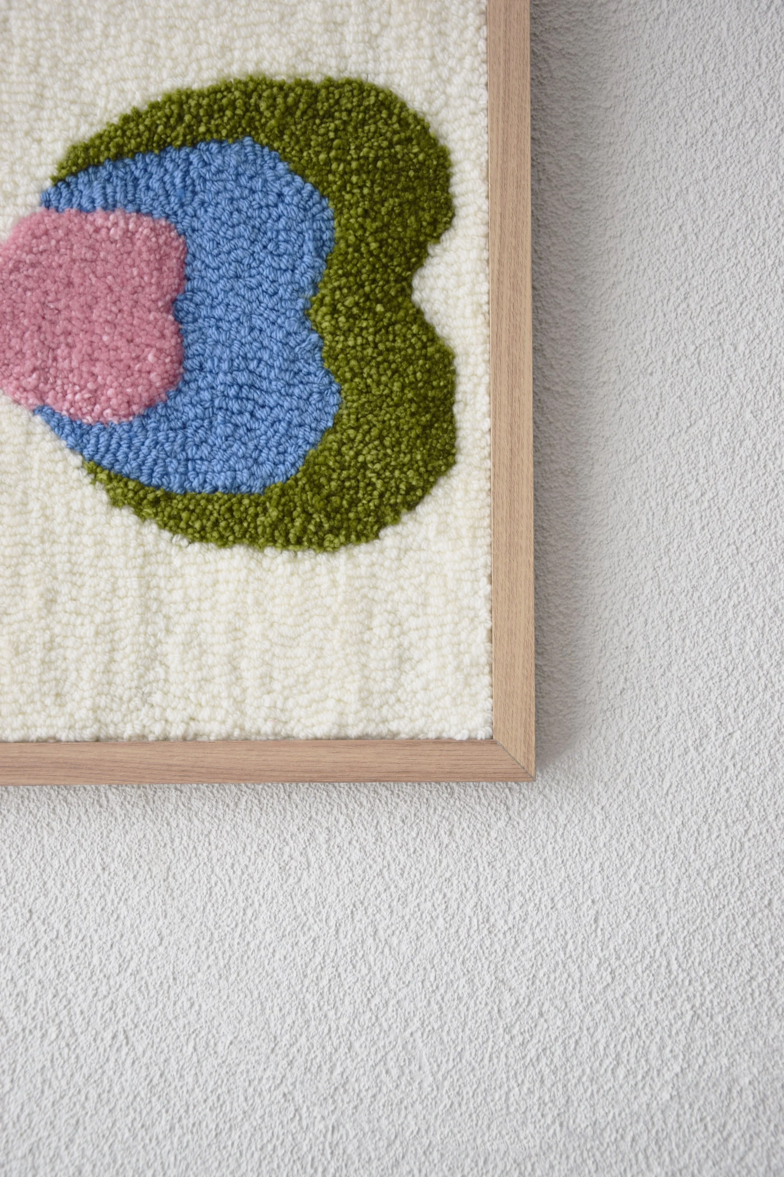 Close-up of a framed, textured textile artwork with abstract shapes in pink, blue, green, and white, mounted on a wall.