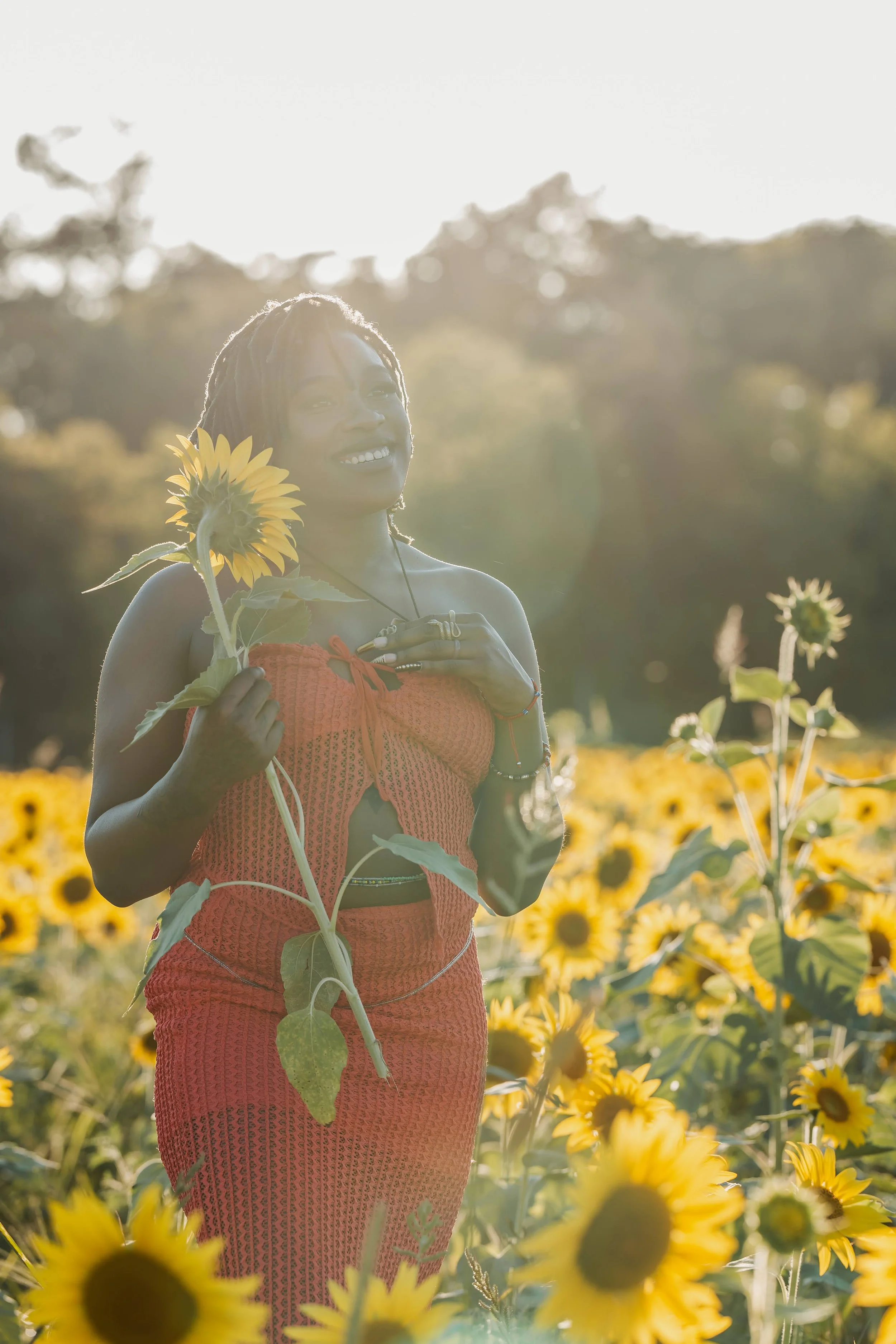 A woman standing in a sunflower field holding a sunflower, smiling, with sunlight shining behind her.