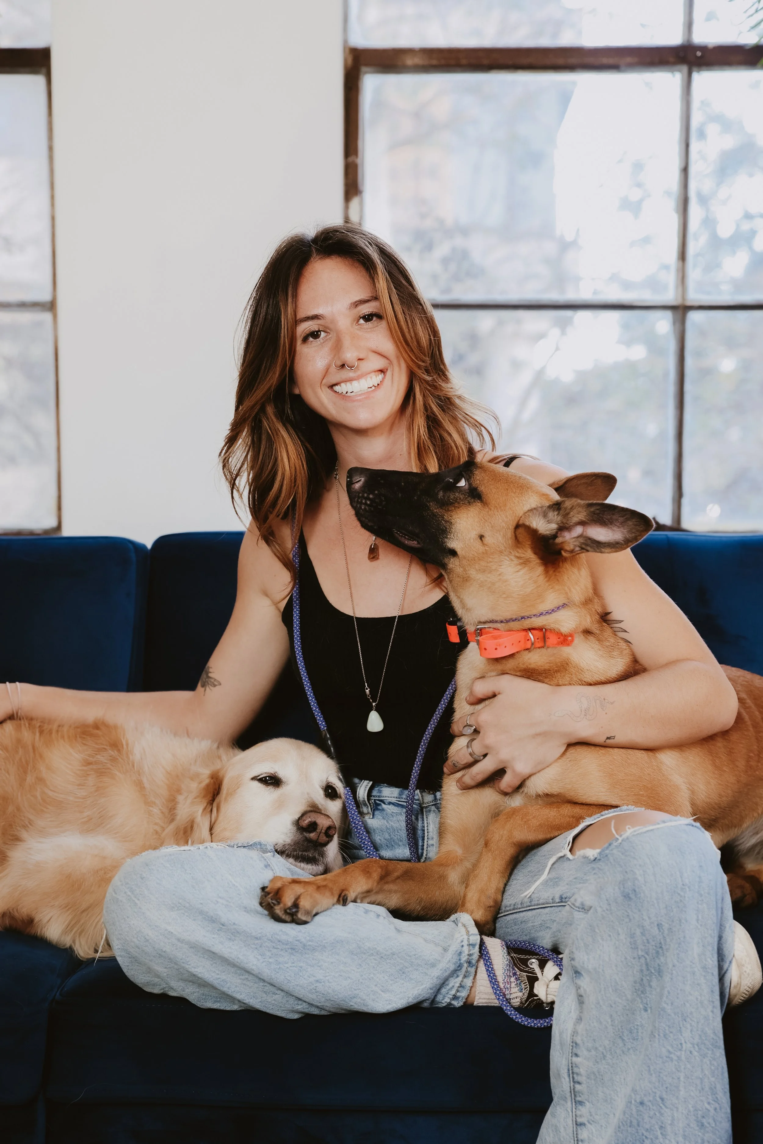 A woman with shoulder-length brown hair, smiling, sitting on a blue couch, holding two dogs; one large tan and black dog with a bright orange collar, and one smaller lighter-colored dog resting its head on her leg.