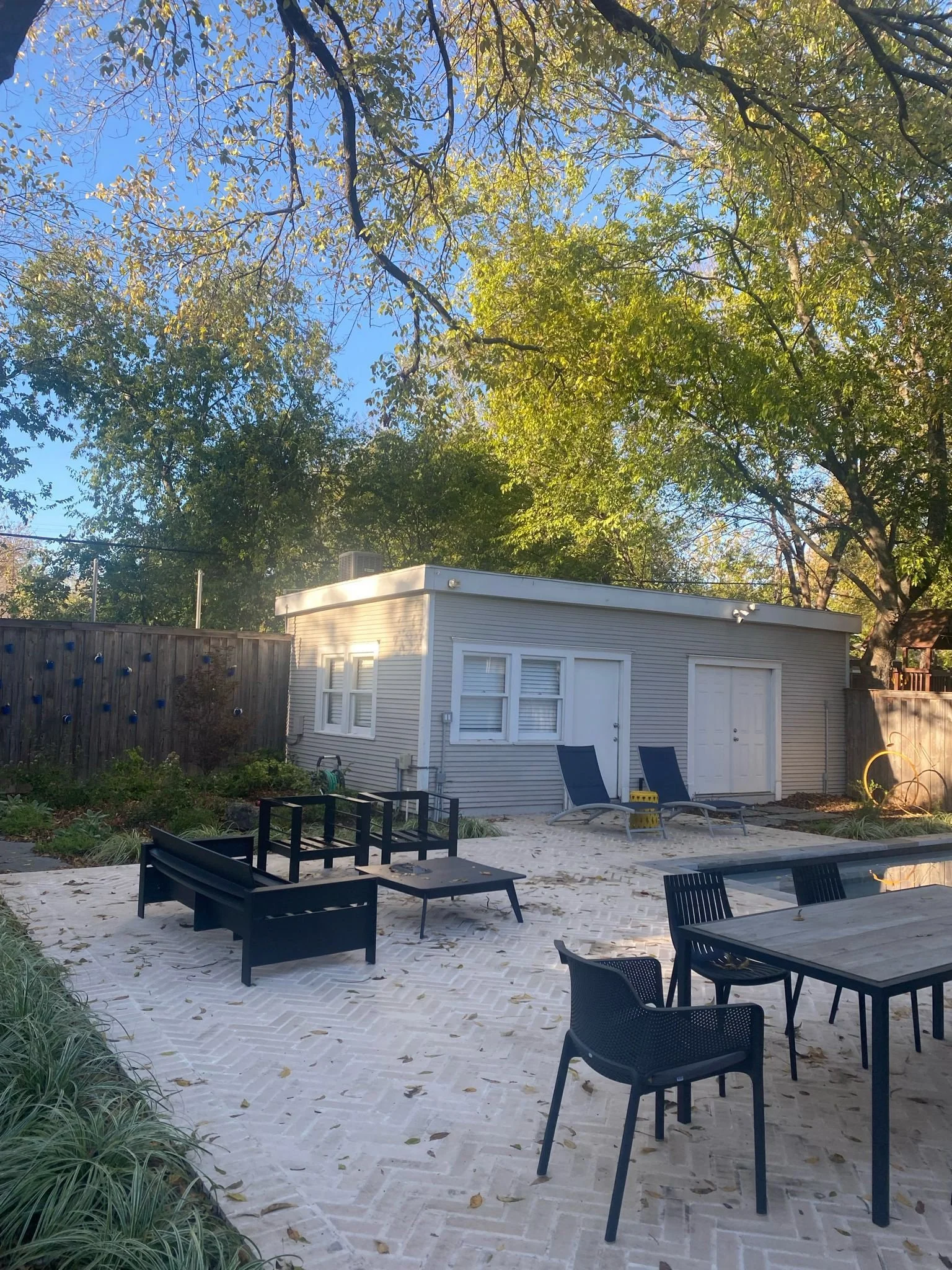 Backyard patio with outdoor furniture, including black chairs, a table, and loungers, near a small modern building surrounded by trees.