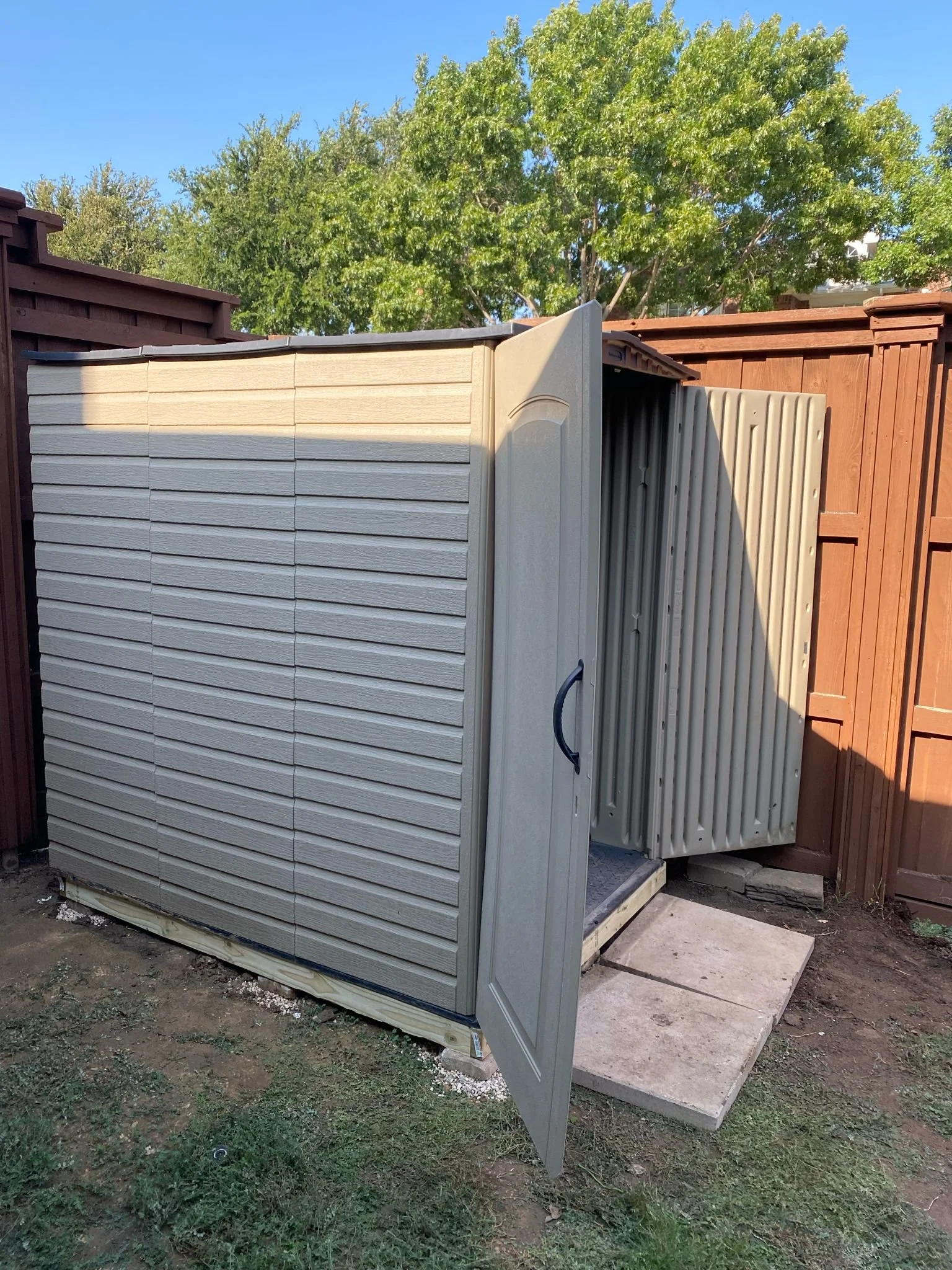 Small outdoor storage shed with open door, situated on concrete slabs in a fenced yard, surrounded by greenery and trees.