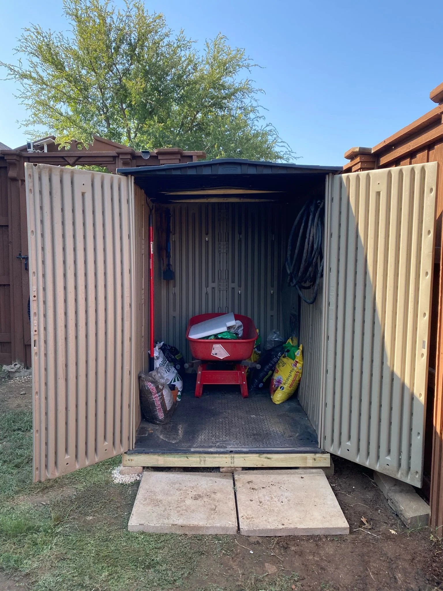 Open metal storage shed with a red wheelbarrow and garden supplies inside.