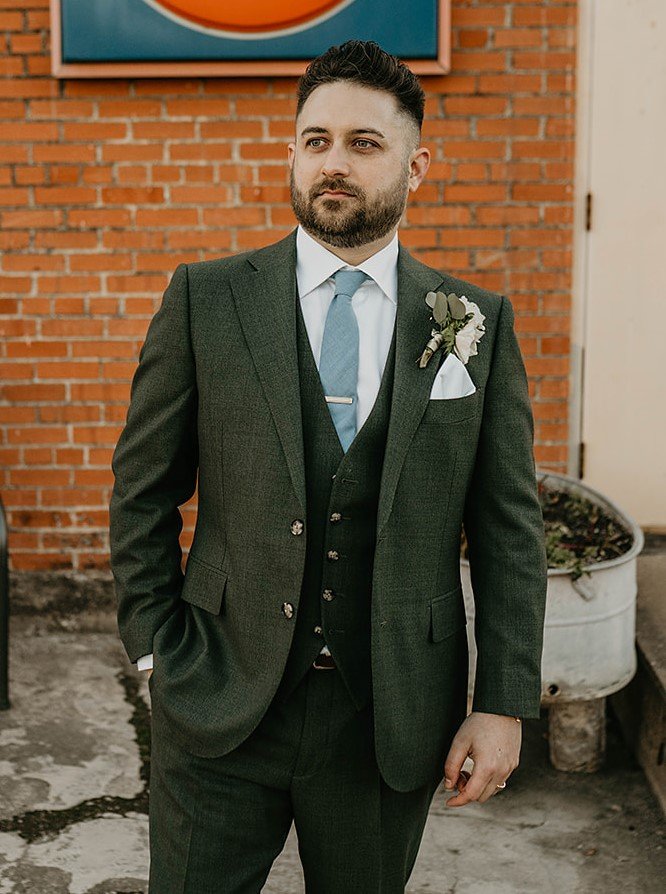 Man in green suit with boutonniere standing against brick wall.