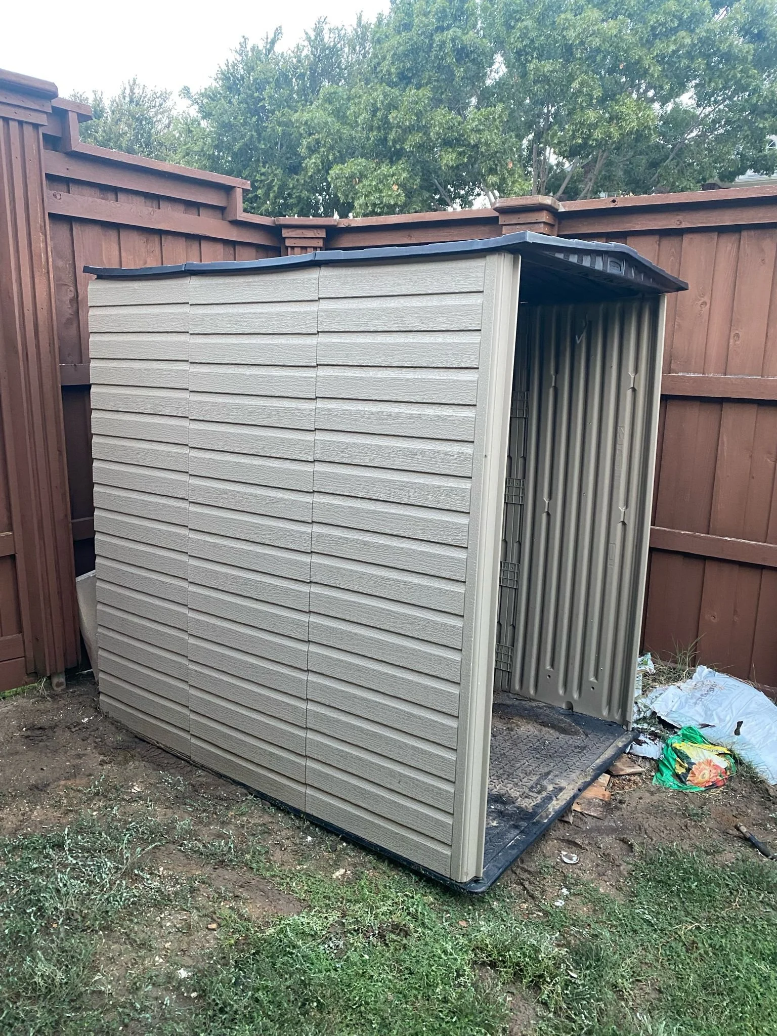 A small, partially open outdoor storage shed next to a wooden fence, with a bag of potting soil on the ground nearby and trees in the background.