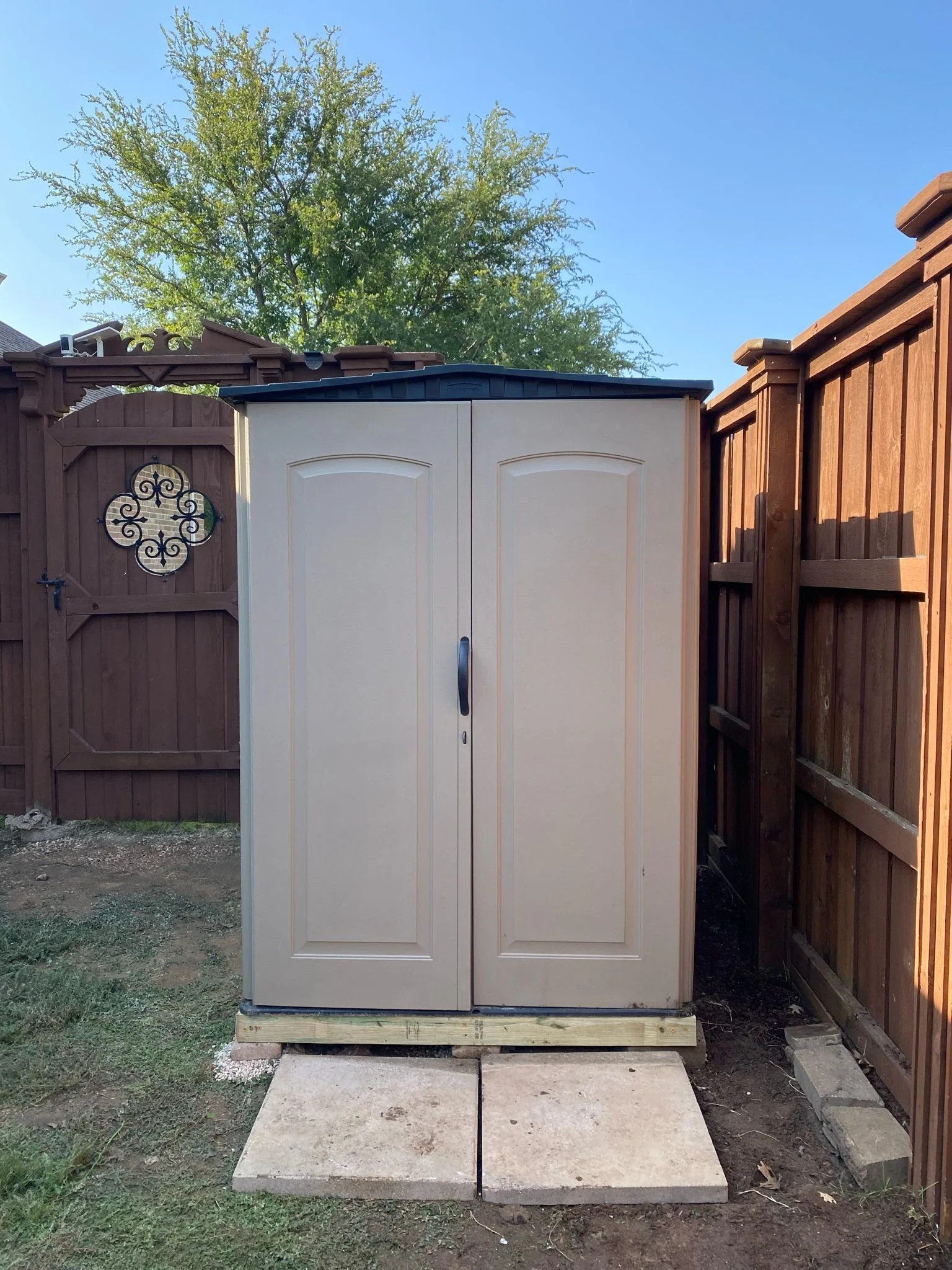 A beige outdoor storage shed with two doors set on concrete slabs in a backyard with a wooden fence and a tree in the background.