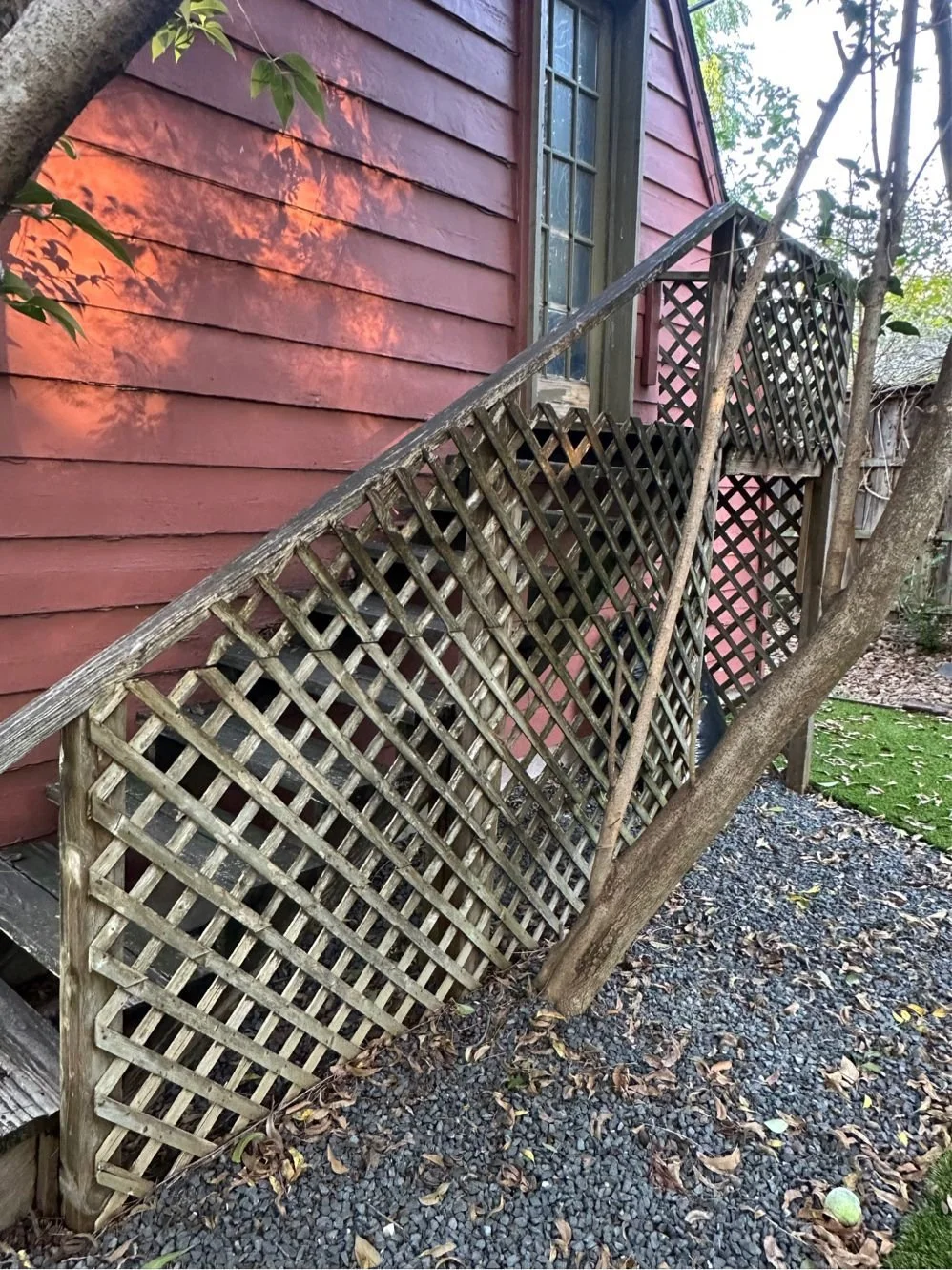 Outdoor wooden staircase with lattice railing attached to a red clapboard house. Deciduous tree nearby with scattered fallen leaves on gravel.