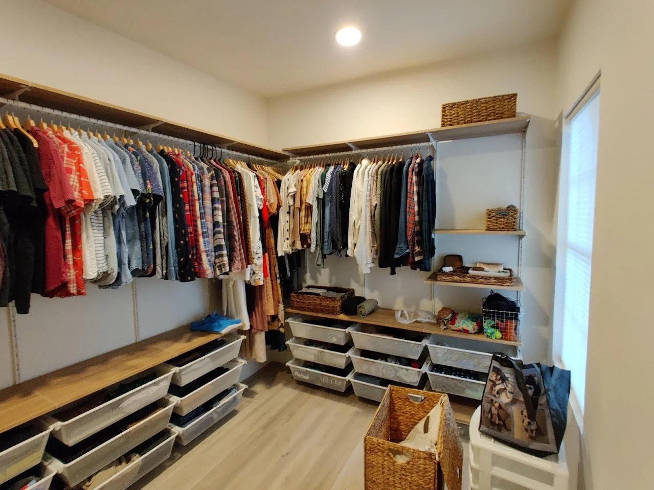 Walk-in closet with neatly organized clothes on hangers, storage baskets on shelves, plastic bins with folded clothes, and a wooden floor.