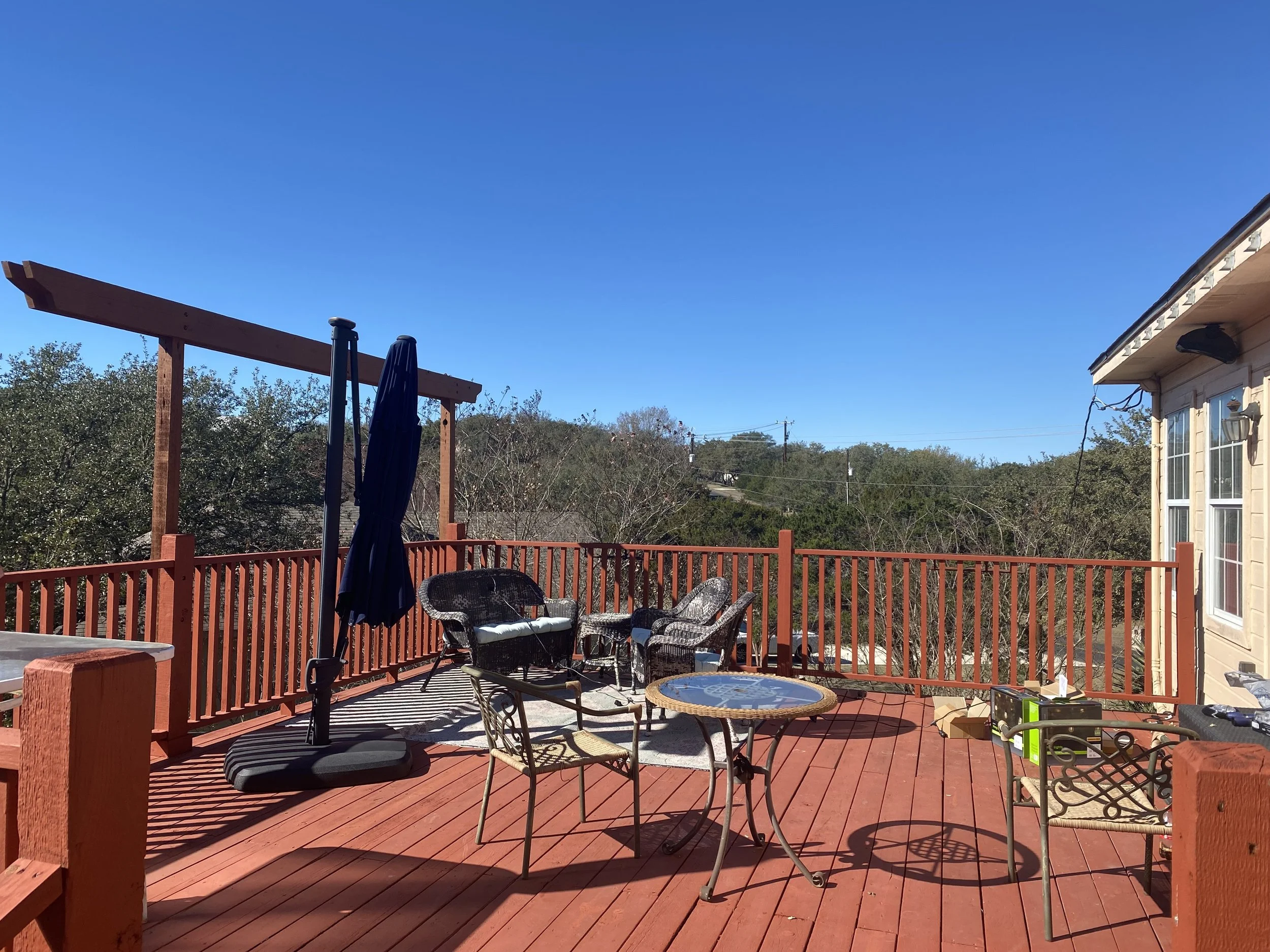 Wooden deck patio with outdoor furniture, enclosed by a railing, and surrounded by trees under a clear blue sky.