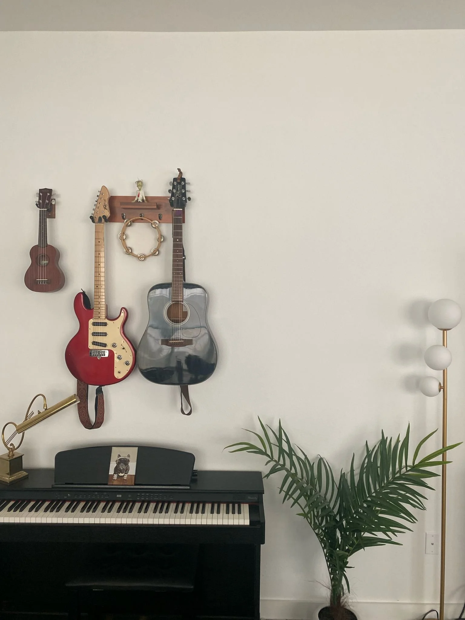 Music room with instruments, including a ukulele, electric guitar, and acoustic guitar mounted on a wall, a digital piano below, and a plant on the floor. A lamp with globe bulbs stands nearby.