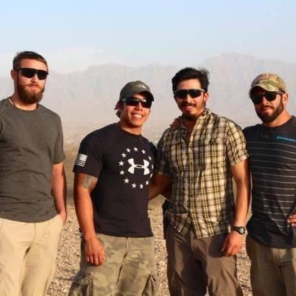Four men wearing casual clothing and sunglasses standing outdoors in a desert landscape during daytime.
