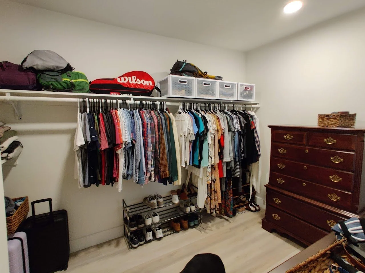 A well-organized closet with hanging clothes, suitcases, a Wilson tennis racket case on the top shelf, and a wooden dresser.