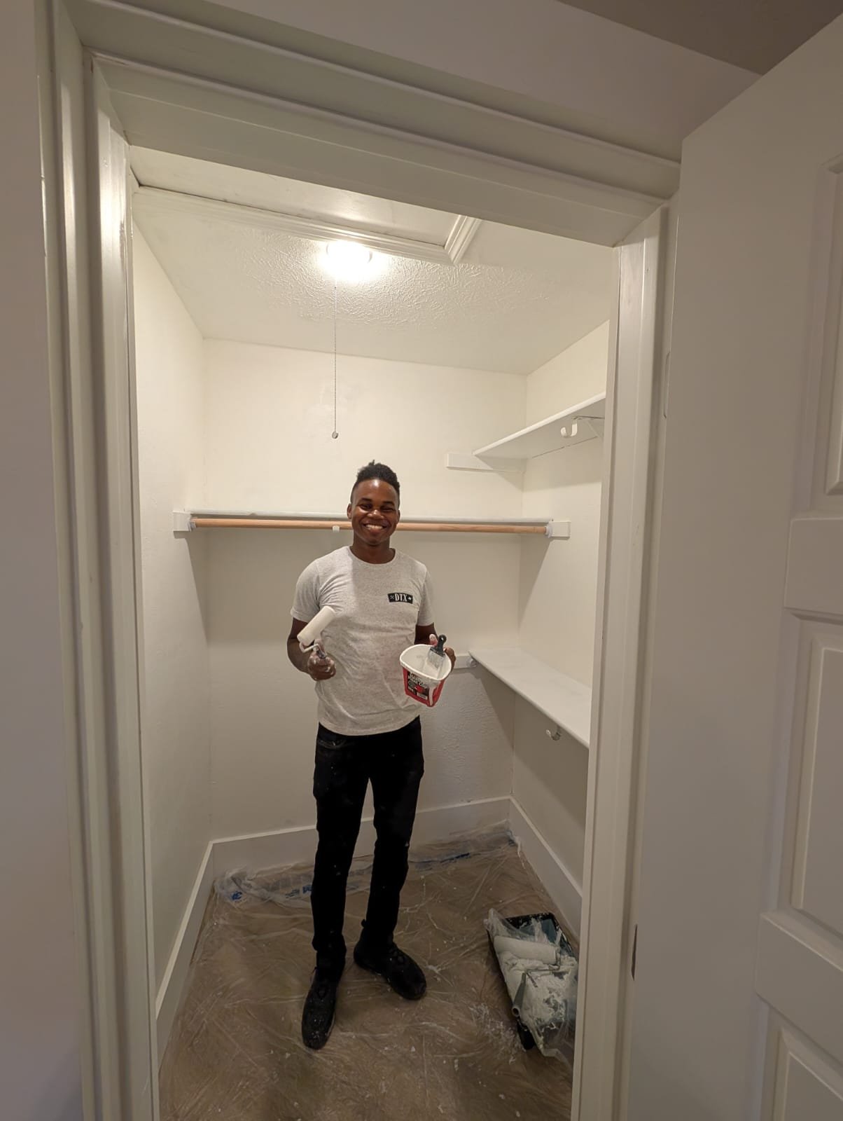 Person standing in a walk-in closet holding painting supplies, including a paint roller and a container, with a protective floor covering and shelves visible.
