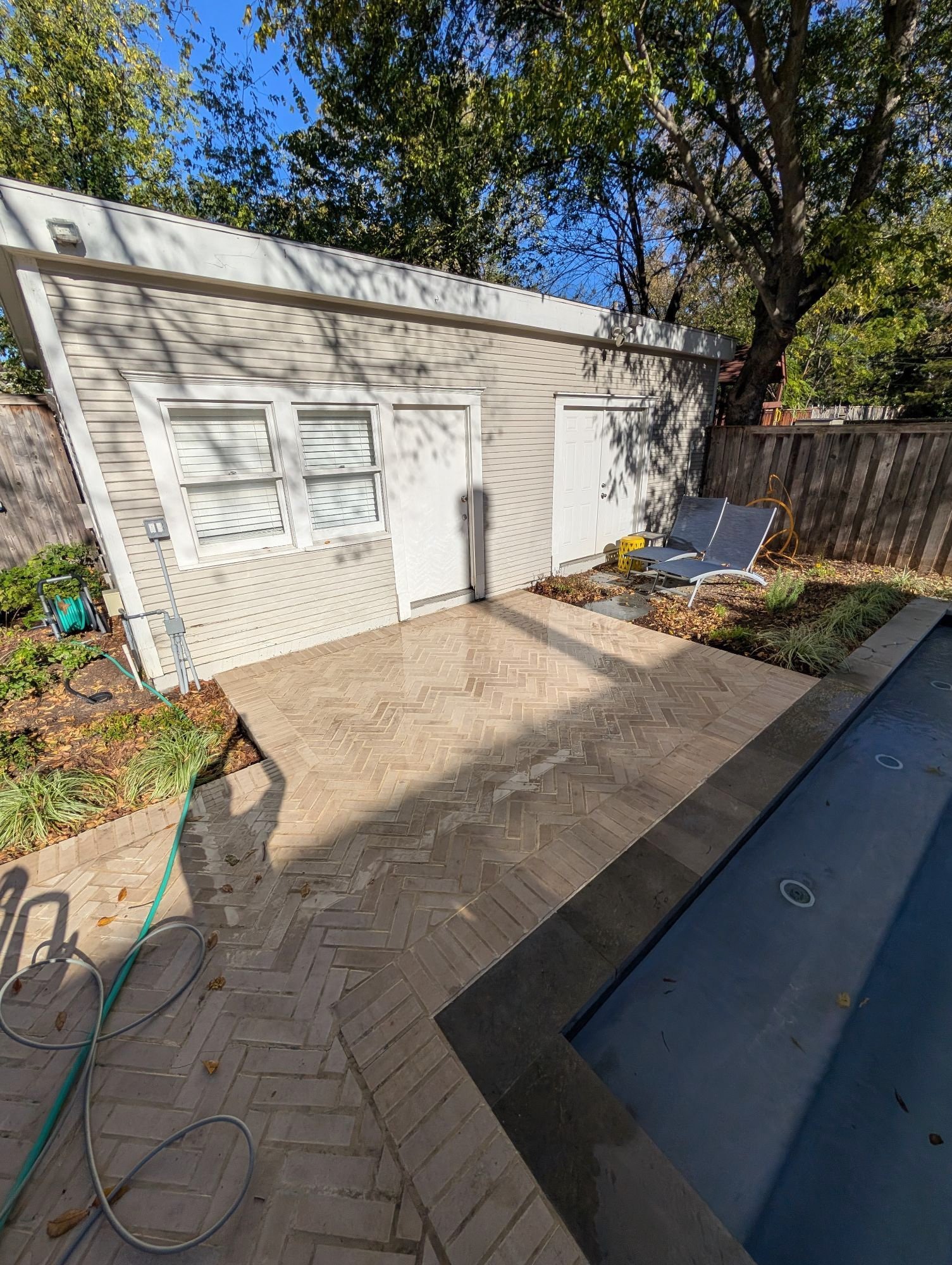 Backyard with a small pool, brick patio, lounge chairs, garden tools, and a storage shed. Trees and a wooden fence are in the background.
