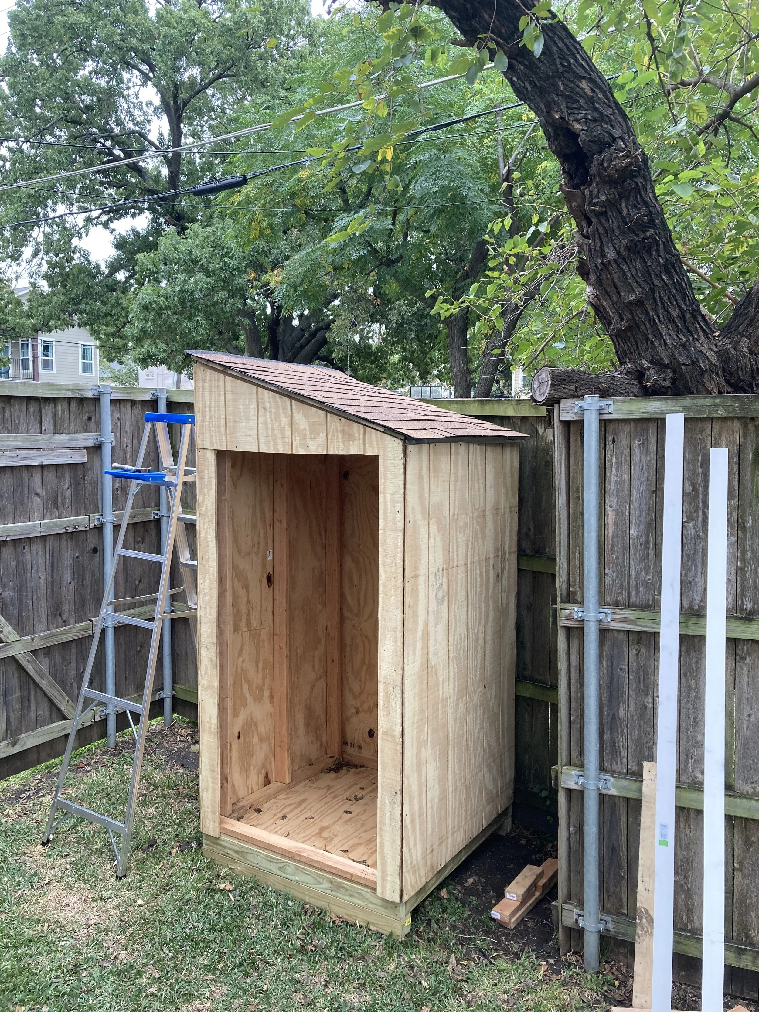 Partially constructed wooden shed with an open front, located beside a fence in a backyard. A ladder and some wooden planks are nearby, with trees and greenery surrounding the area.