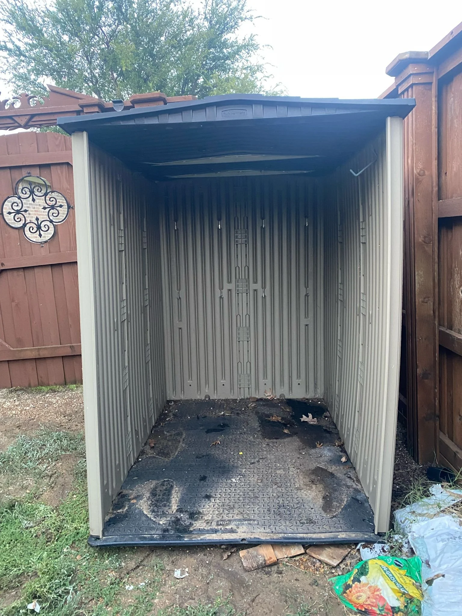Empty plastic garden shed in a backyard with a wooden fence and some garden tools and bags nearby.